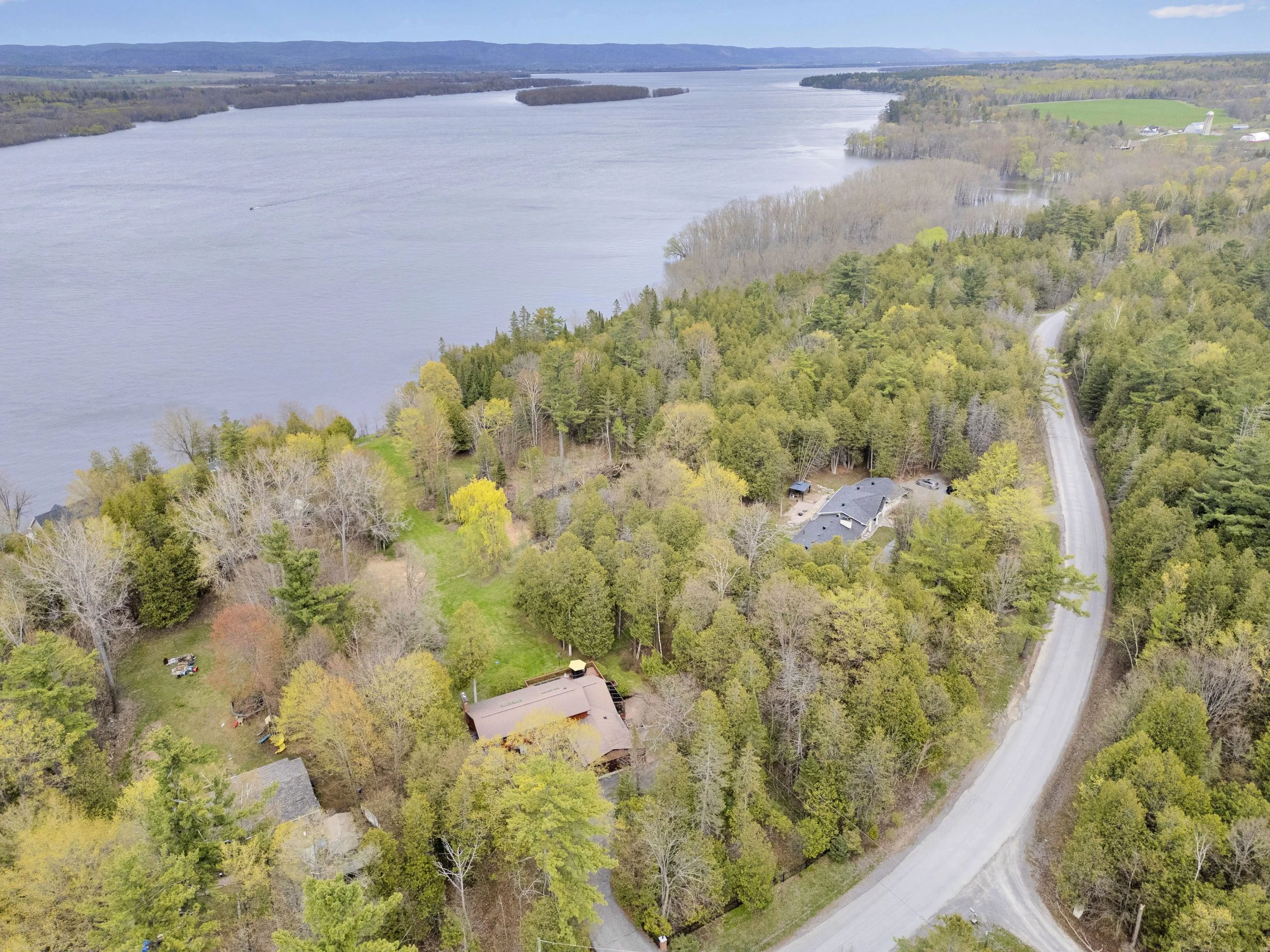 NVISION3D.ca Real Estate Media Ottawa  Aerial view of a large river with boats, surrounded by green trees, farmland, and a few houses, with mountains in the distance under a partly cloudy sky.