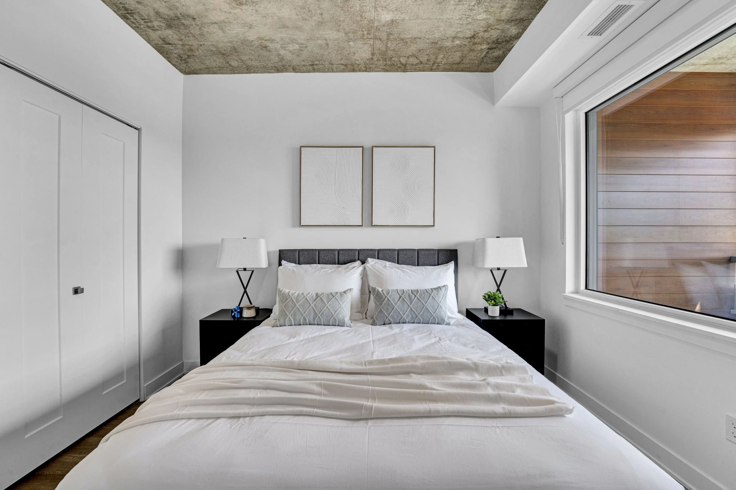Modern bedroom with a bed, white bedding, two pillows, black nightstands with lamps, a small plant, two framed abstract art pieces on the wall, a large window showing a wooden exterior, a ceiling with a concrete finish, and white walls.