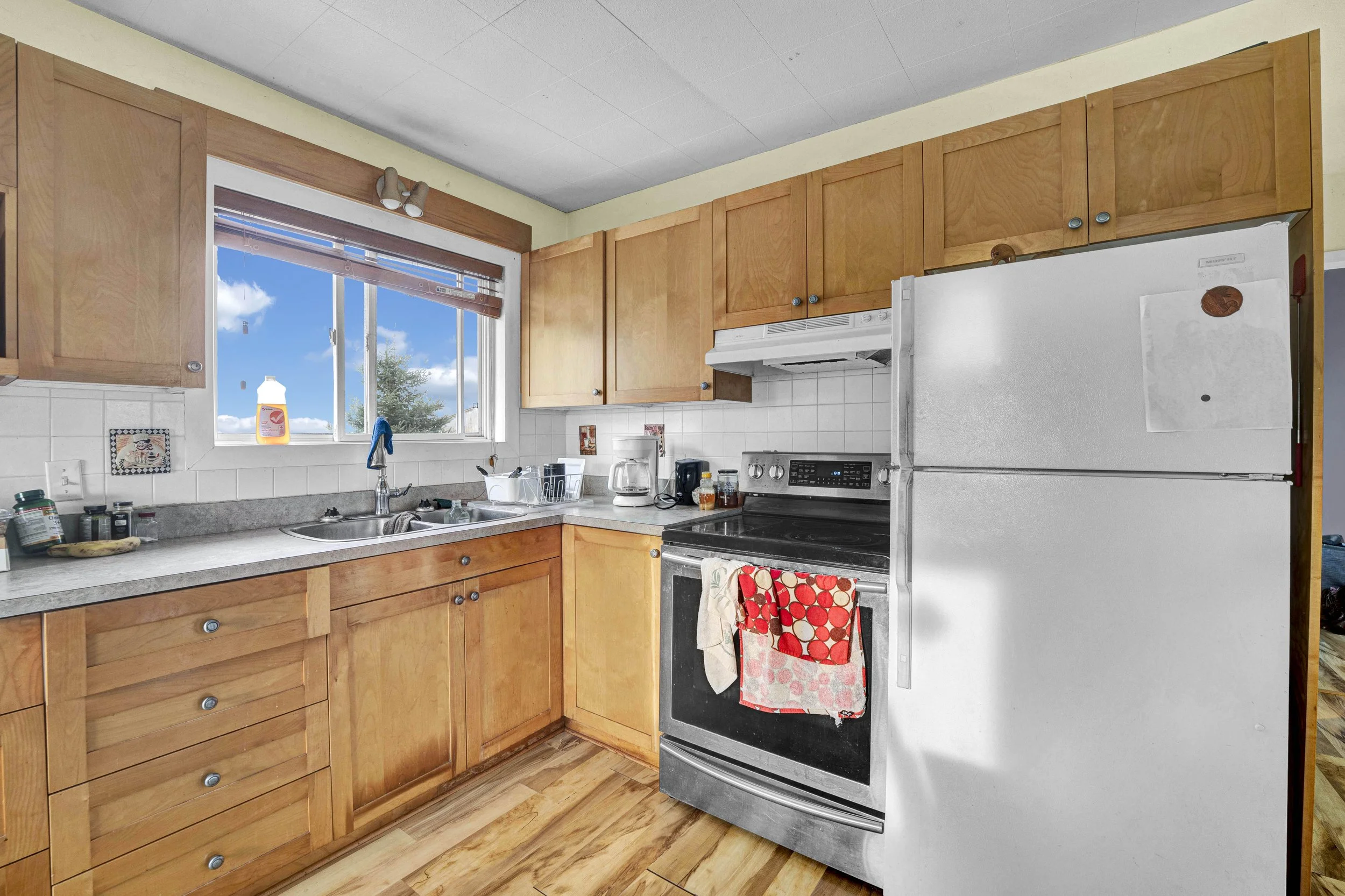 Kitchen with wooden cabinets, a window with a blue sky and clouds, a white refrigerator, a stove with colorful towels hanging, and various kitchen items on the counter.