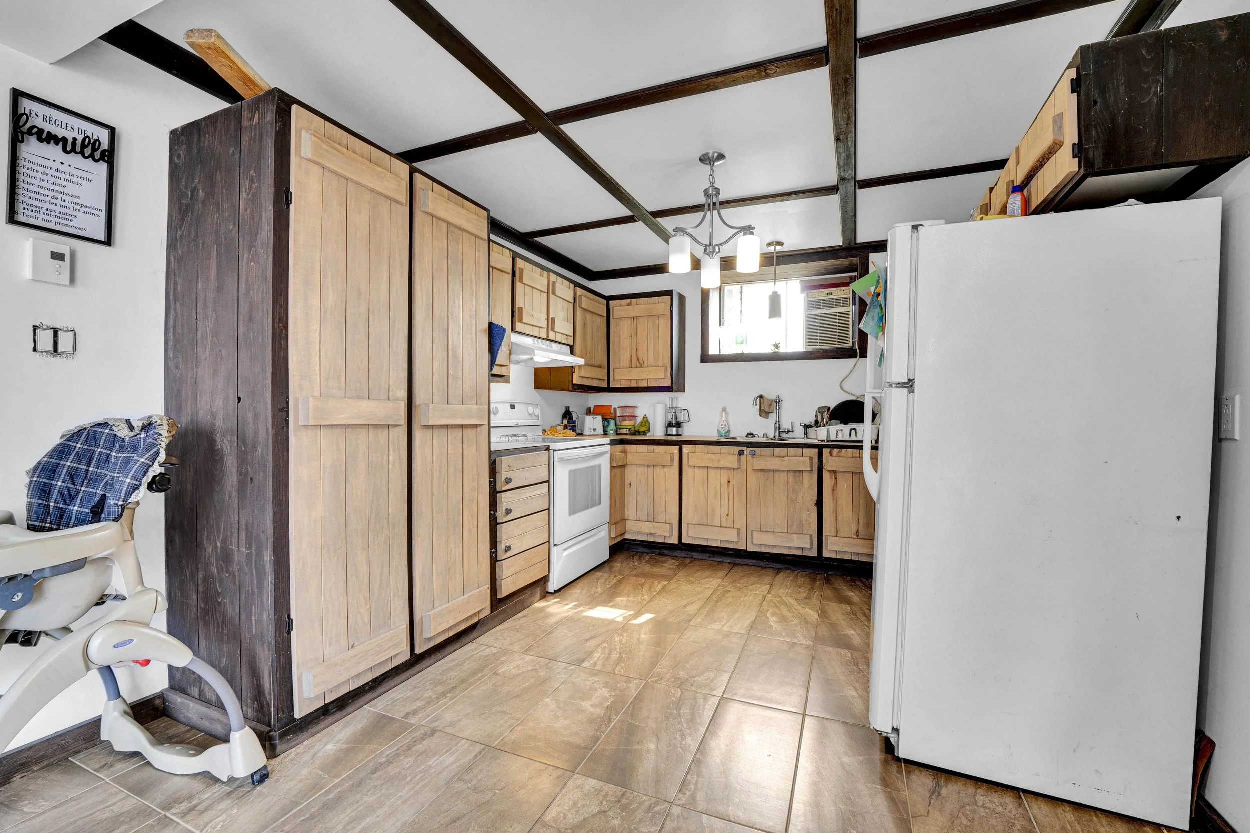 A kitchen with wooden cabinets, a white stove, and a window air conditioning unit in the corner. There's a baby high chair with a blue checkered blanket on it and a framed quote on the wall in French.