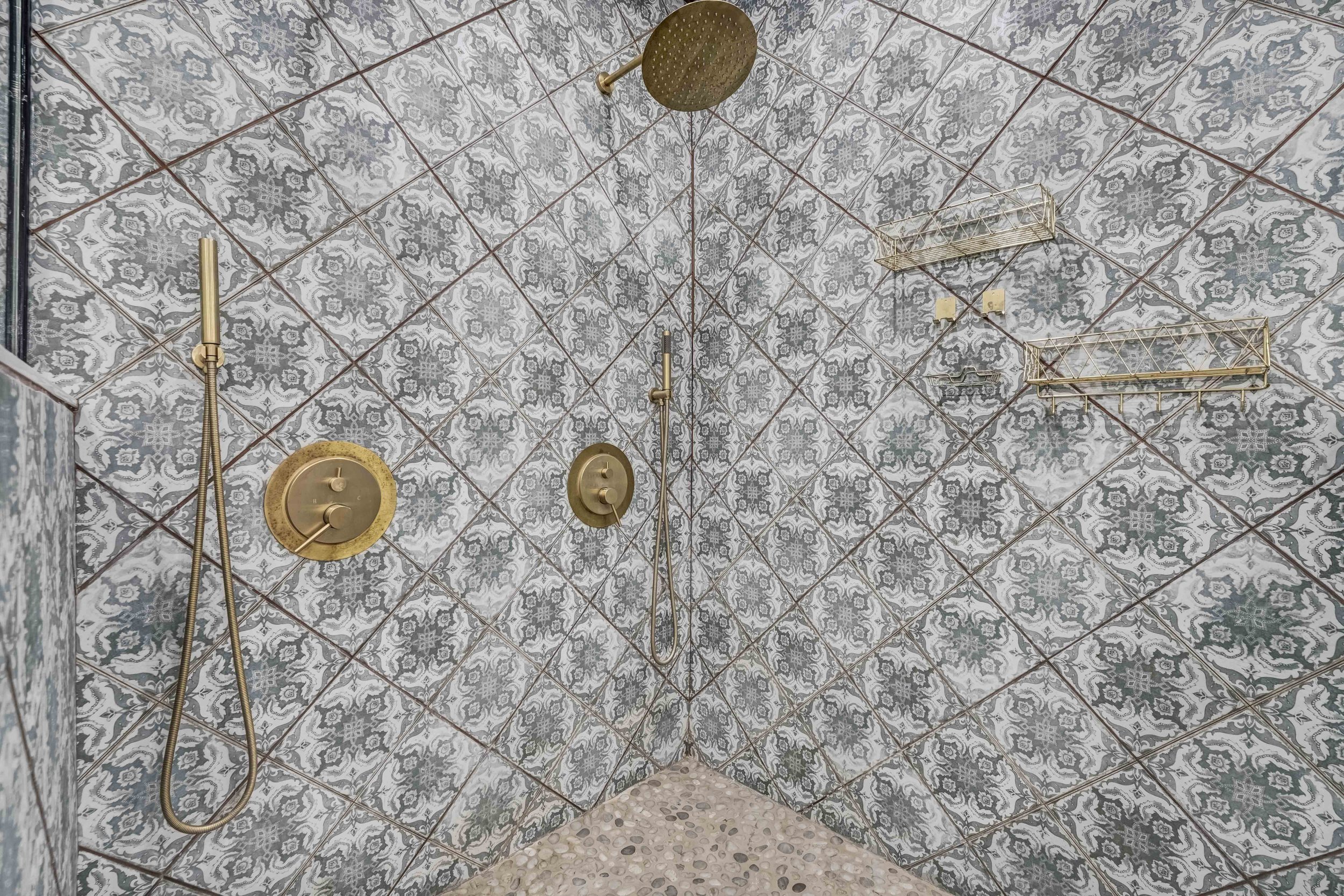 A walk-in shower with patterned gray and white tiles, a round brass showerhead, a brass hand shower, a brass temperature control, and brass shelves mounted on the wall. Ottawa Real Estate Photography