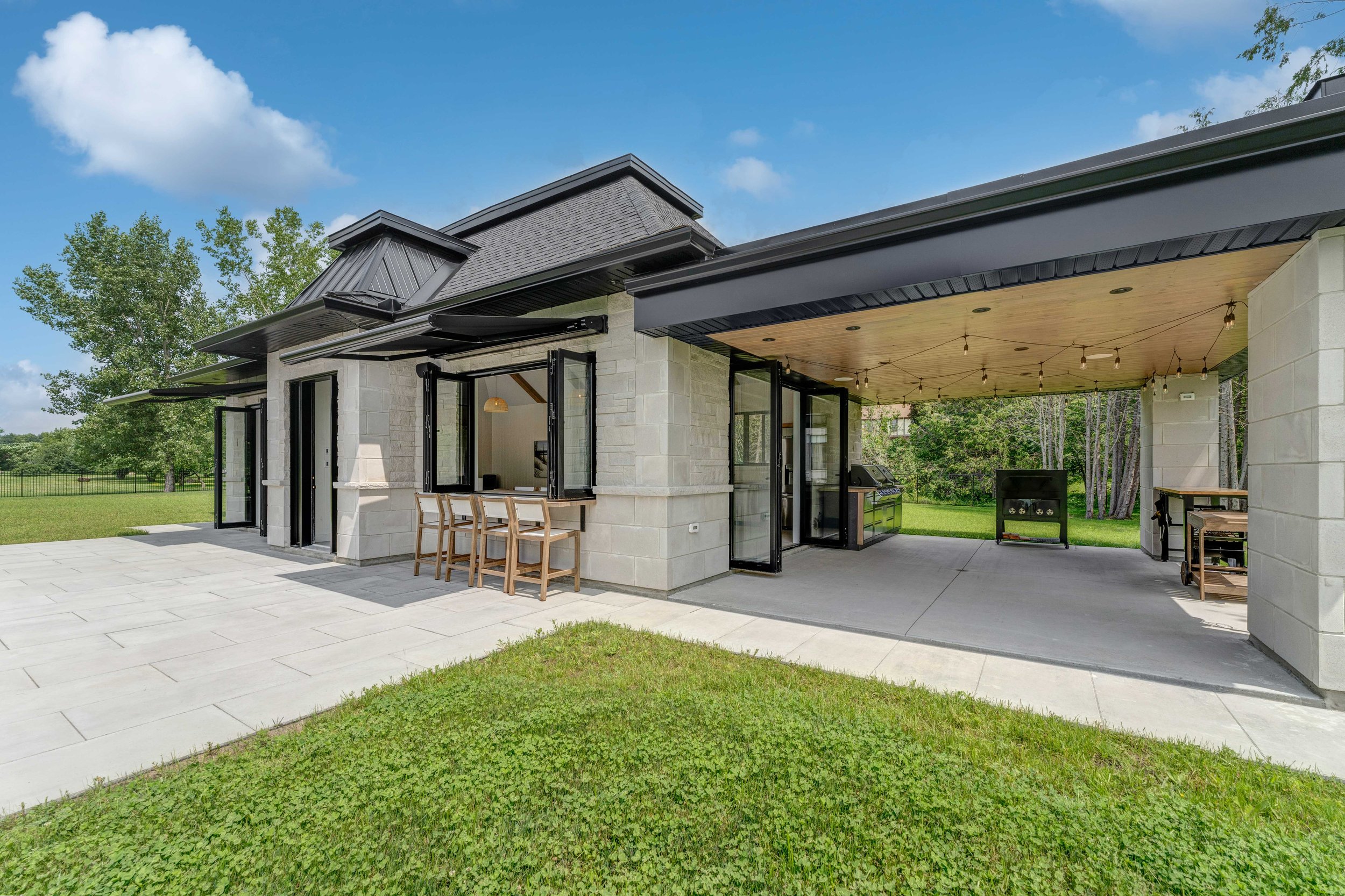 Modern outdoor patio with stone facade, seating area, and grill under a wooden ceiling. Large glass doors and lush green lawn visible.