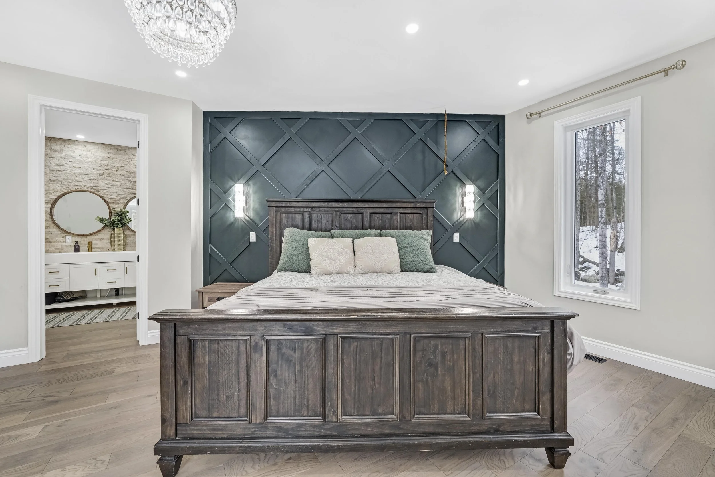 photographie immobiliere outaouis  Bedroom with a wooden bed frame and a dark teal accent wall behind it, decorative pillows, large window showing snow outside, doorway to a bathroom with a mirror and vase inside.