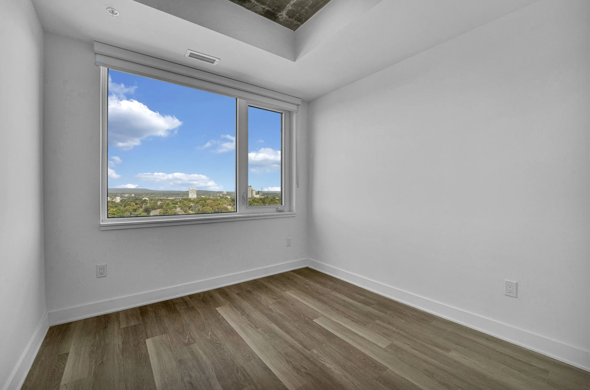 Empty room with white walls, hardwood flooring, a large window showing a cityscape and blue sky with clouds, and a ceiling with mixed materials.
