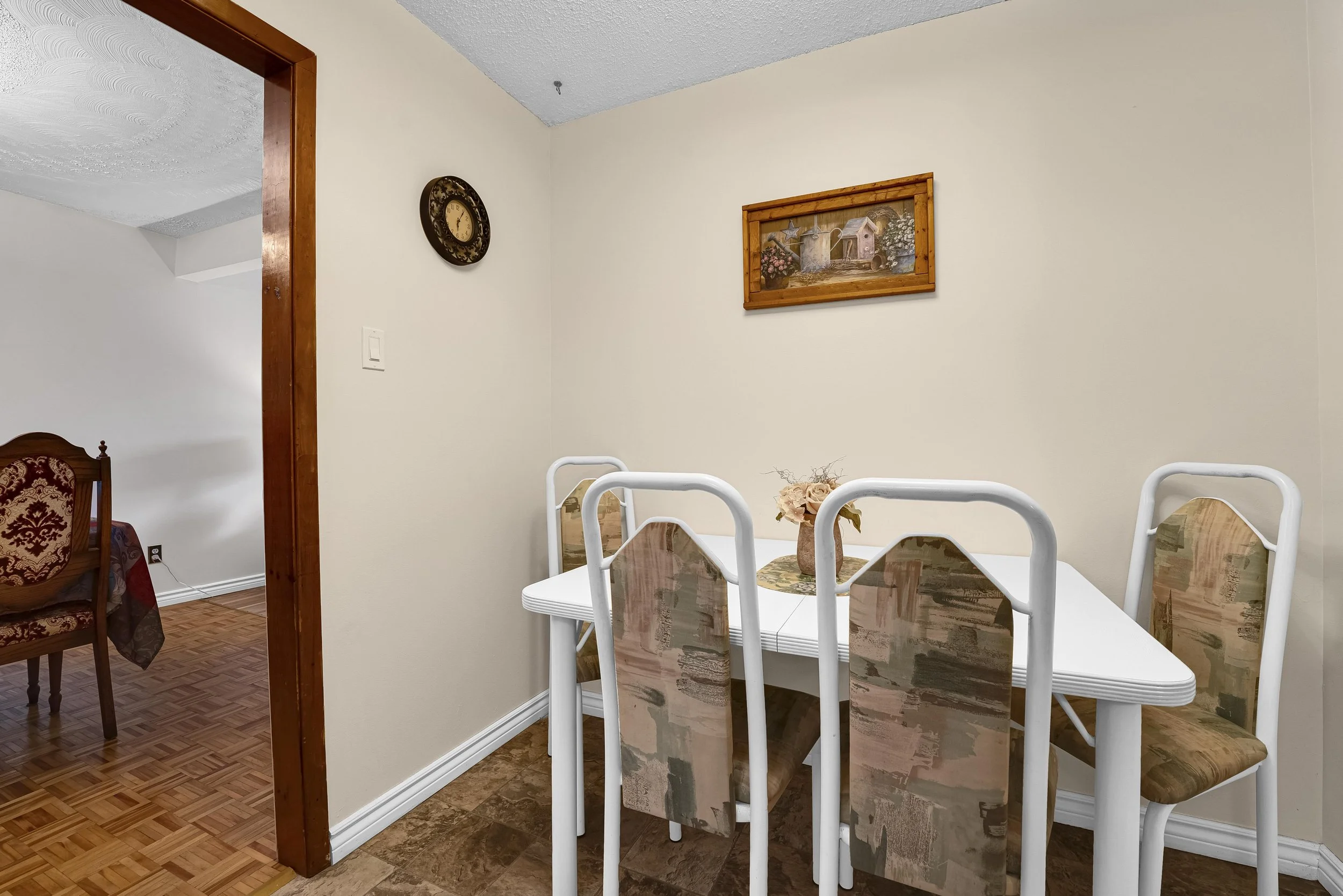 Ottawa Real Estate Photographer Dining room with a white table, several chairs, and flower centerpiece. The wall has a picture of a garden scene and a round clock. There's a wooden doorframe and hardwood flooring.