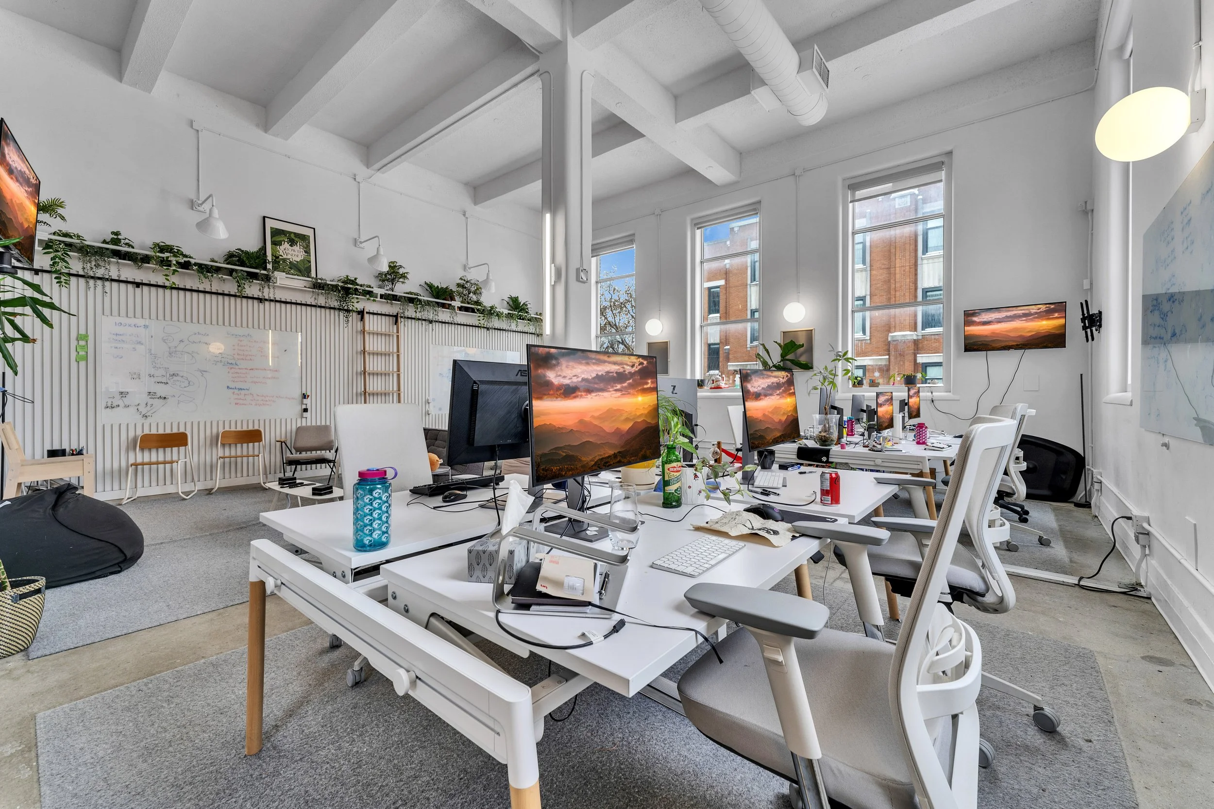 Modern open-plan office with desks, computers, plants, and large windows letting in natural light.