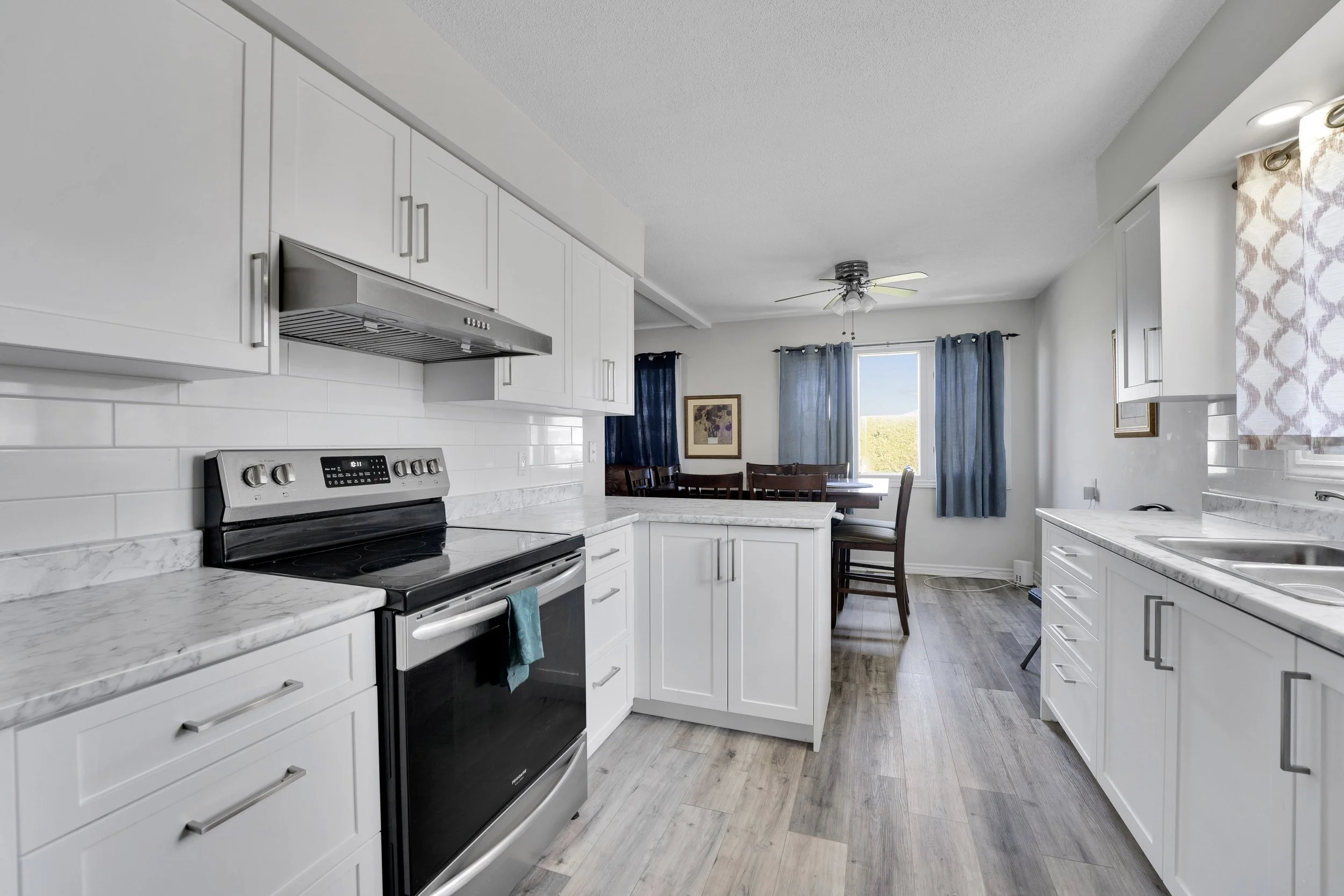 Ottawa Real Estate Media Services  A bright kitchen featuring white cabinets with silver handles, a marble countertop, stainless steel oven and range, white subway tile backsplash, and a window with blue curtains.