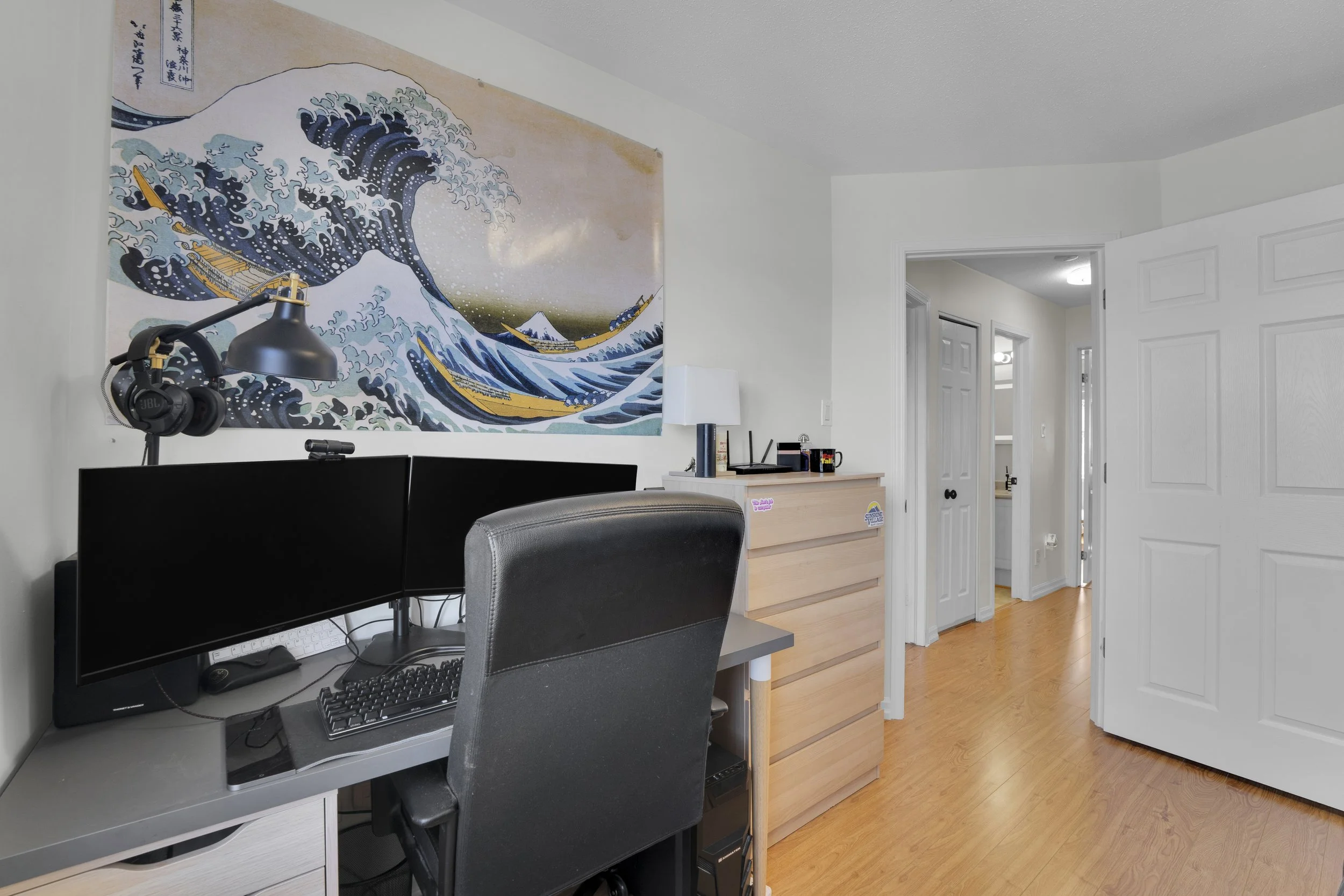 NVISIONPart of a home office featuring a large wall hanging, a computer setup with dual monitors, a keyboard, headphones on a stand, a black office chair, a wooden dresser, and a door leading to a hallway with light-colored walls and wooden flooring.