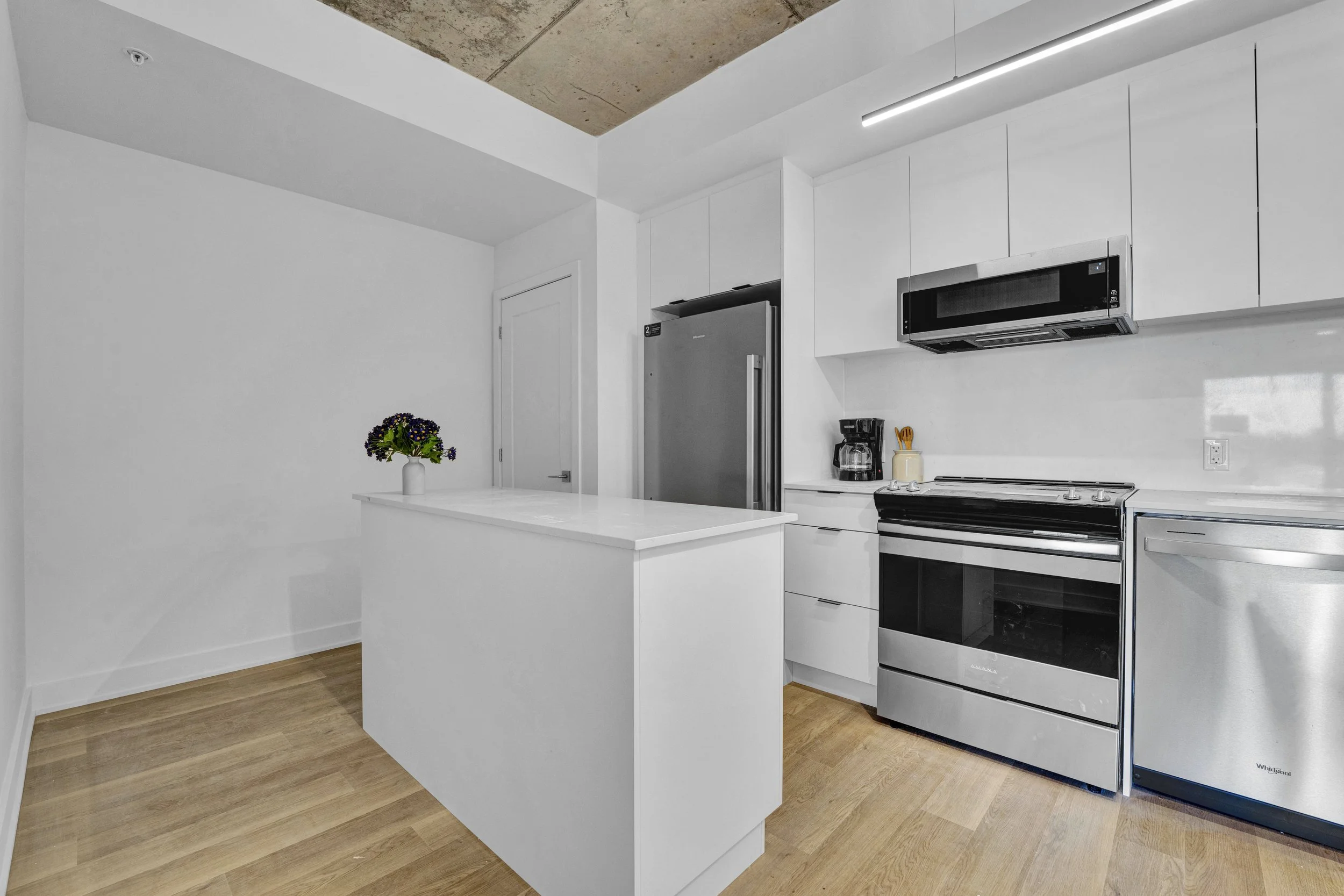 Ottawa Real Estate Media Services  Modern minimalist kitchen with white cabinets, stainless steel appliances, wooden floor, and a small island with a vase of flowers.
