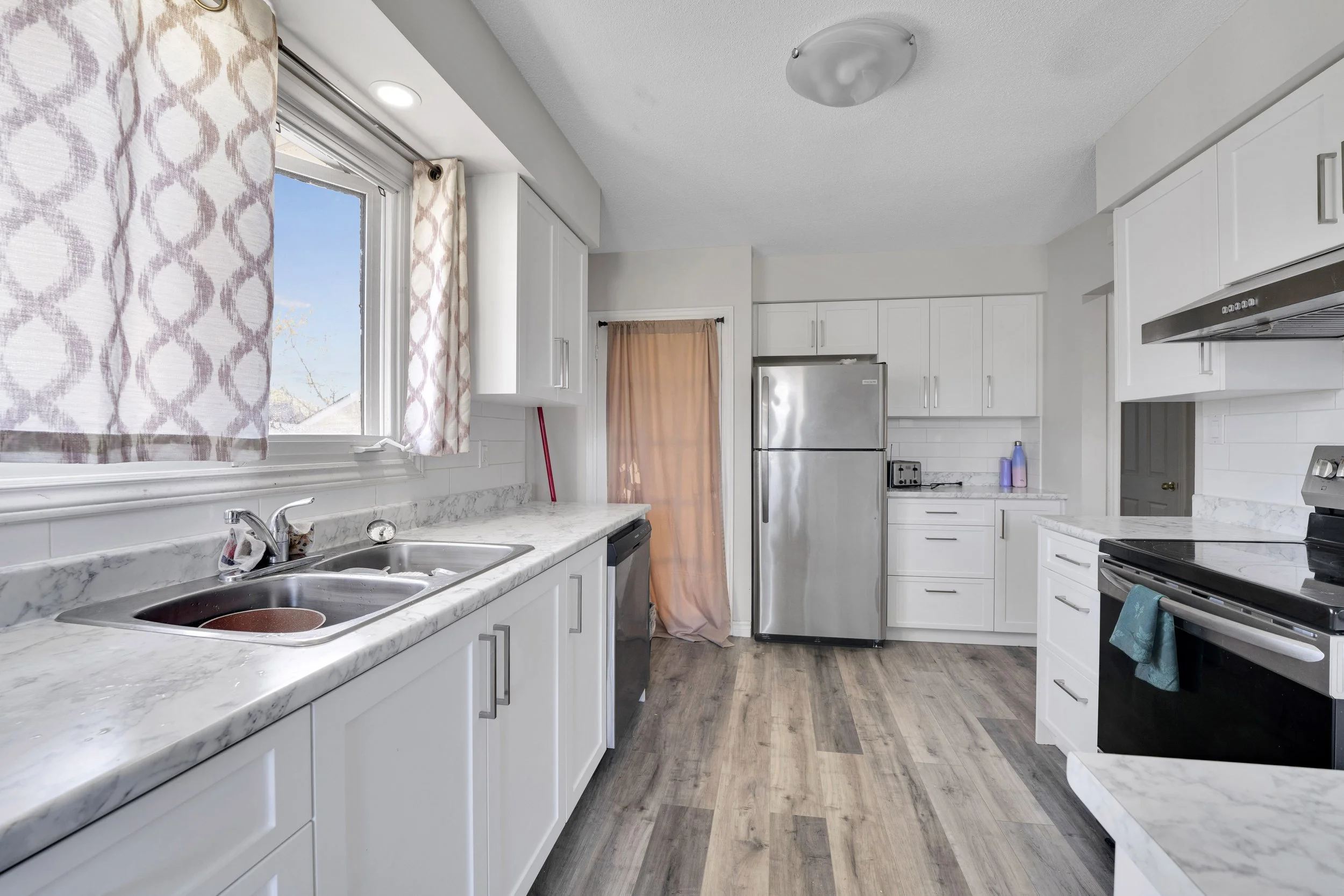 Ottawa Real Estate Media Services  A clean modern kitchen with white cabinets, marble countertops, a stainless steel refrigerator, and a stove. There is a window with patterned curtains, a sink, and a wooden floor.