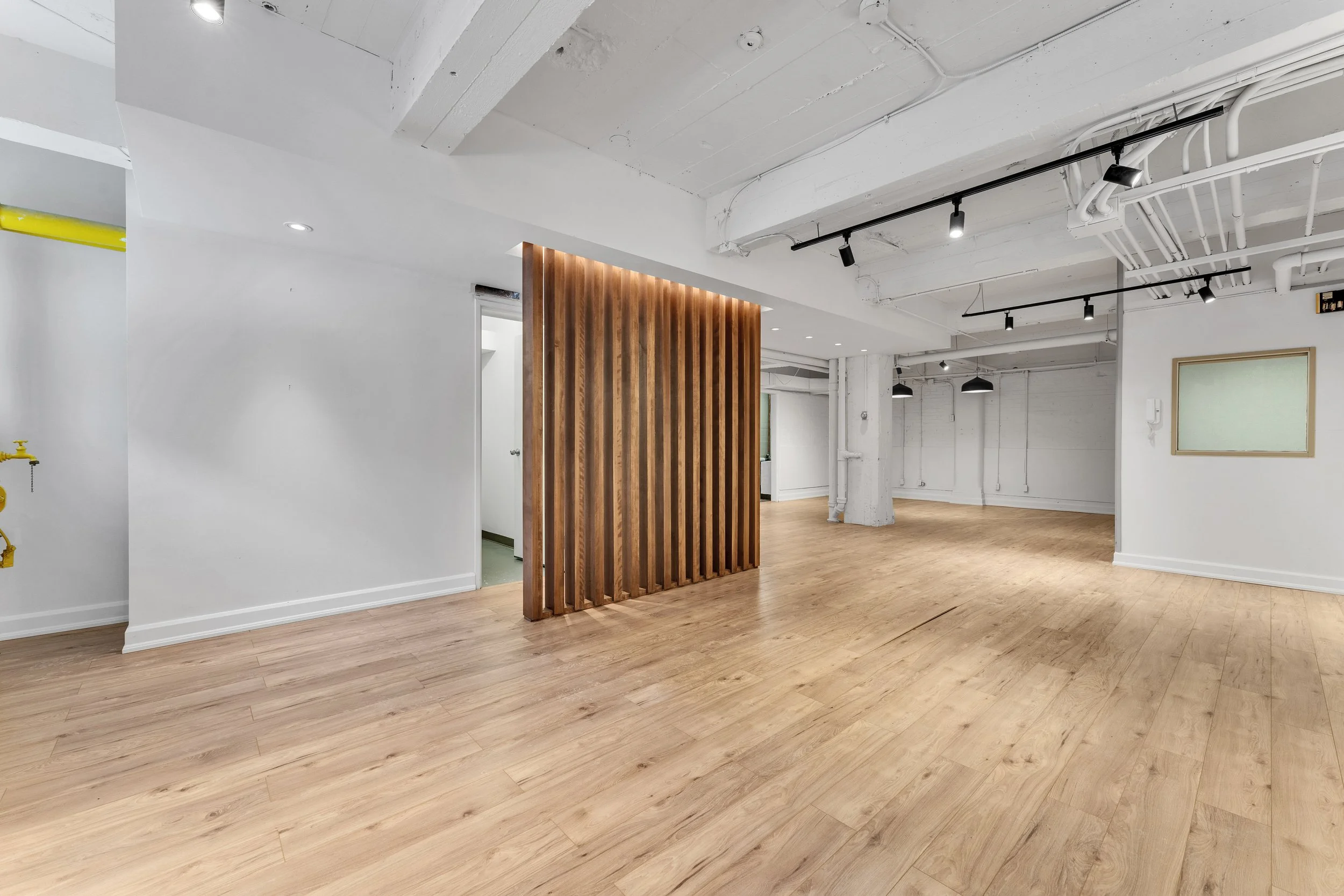 Empty modern interior space with white walls, wooden flooring, exposed ceiling pipes, and a vertical wooden slat partition.