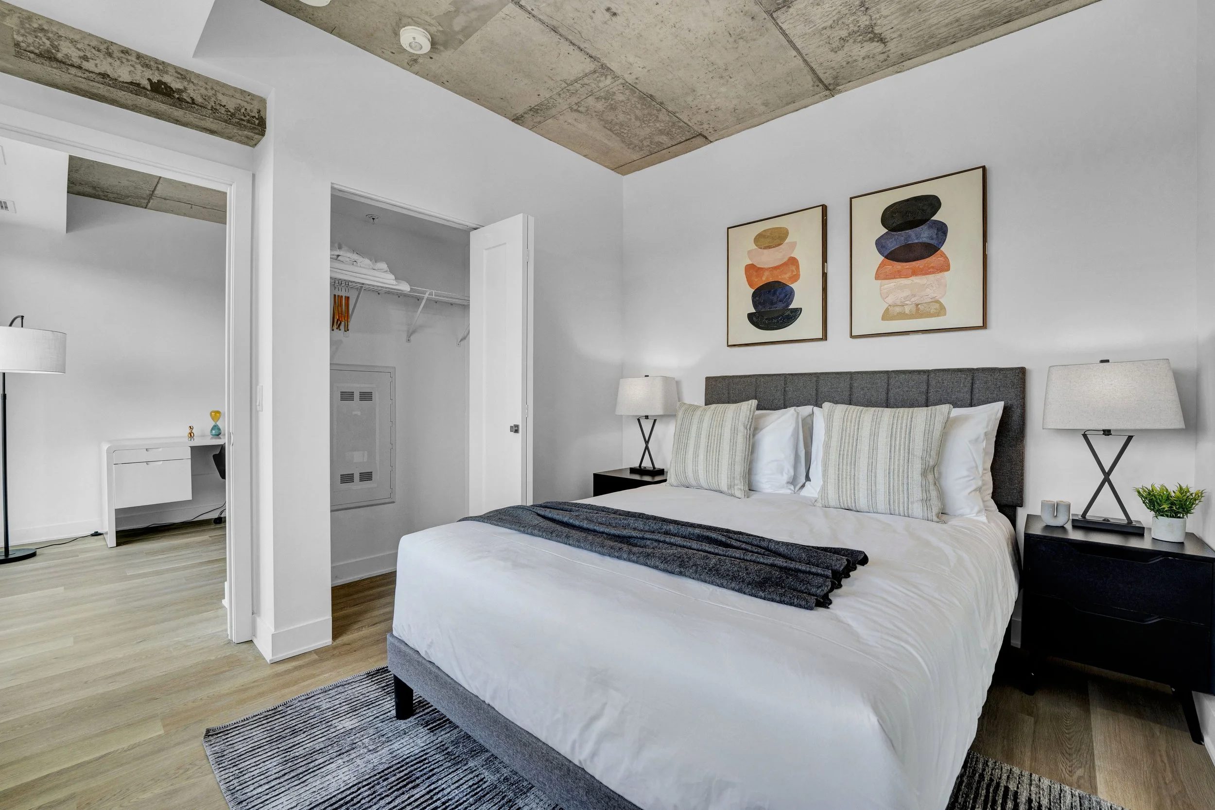 A modern bedroom with a bed, gray upholstered headboard, two nightstands with lamps, abstract artwork above the bed, and a corner with a small writing desk and a lamp. The ceiling has exposed concrete, and the floor is hardwood. There is an open clos