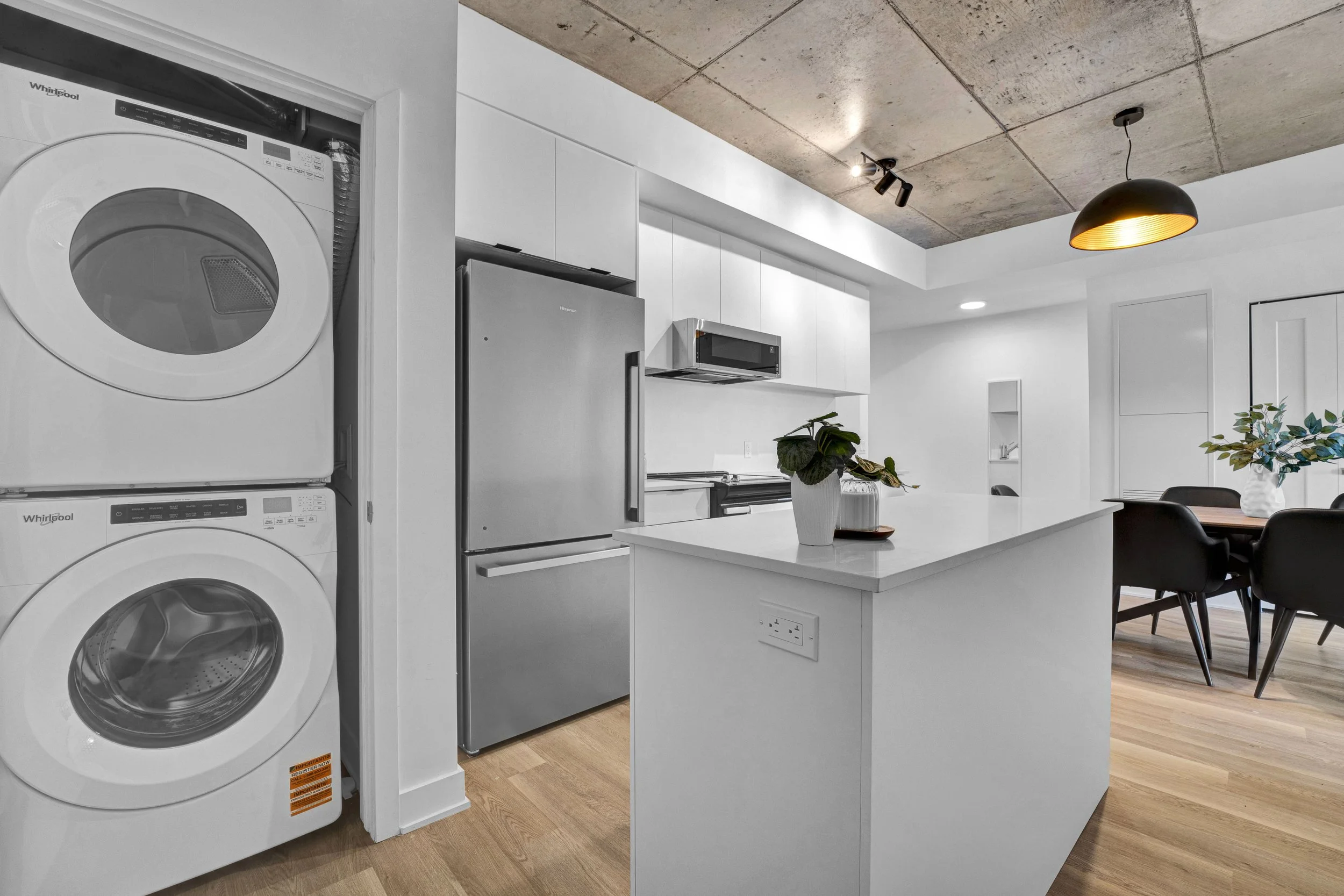 Ottawa Real Estate Media Services  Modern kitchen with white cabinetry, a stainless steel refrigerator, microwave, and a white island with potted plants. Laundry appliances are stacked in a closet next to the kitchen.