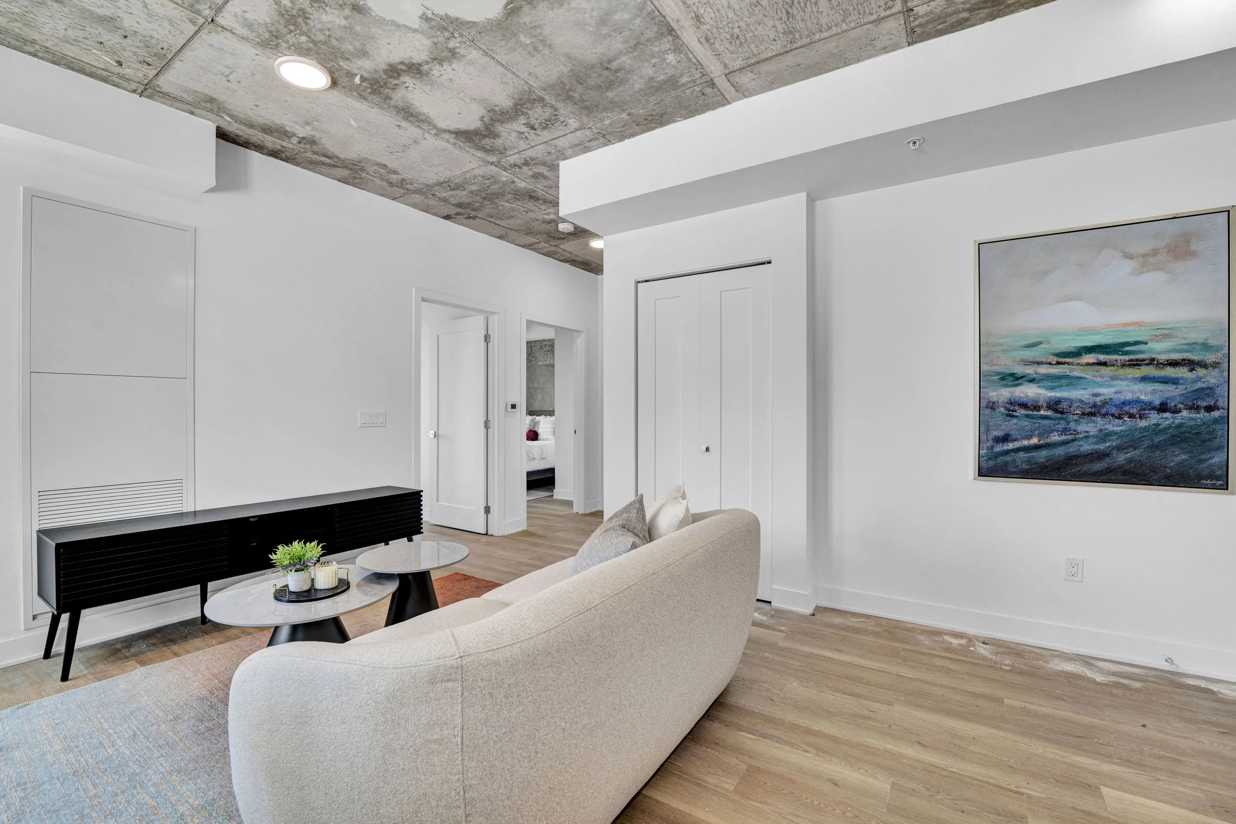 Ottawa Real Estate Photographer Modern living room with white walls, wood flooring, a beige sofa, black coffee table, colorful abstract painting on the wall, and a view into a bedroom with a bed and red accents.