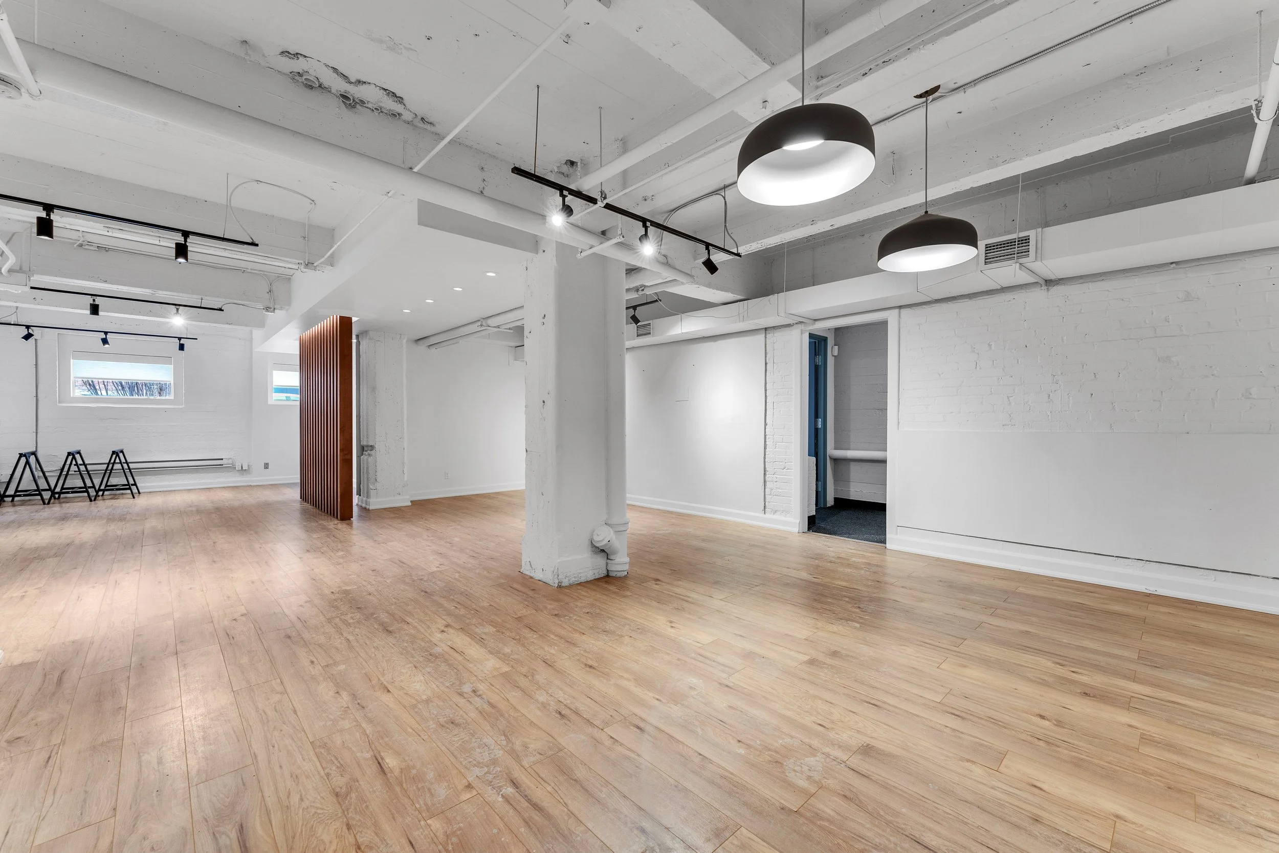 photographie immobiliere outaouis  Empty modern commercial space with white walls, exposed ceiling pipes, wooden flooring, and black pendant lights.