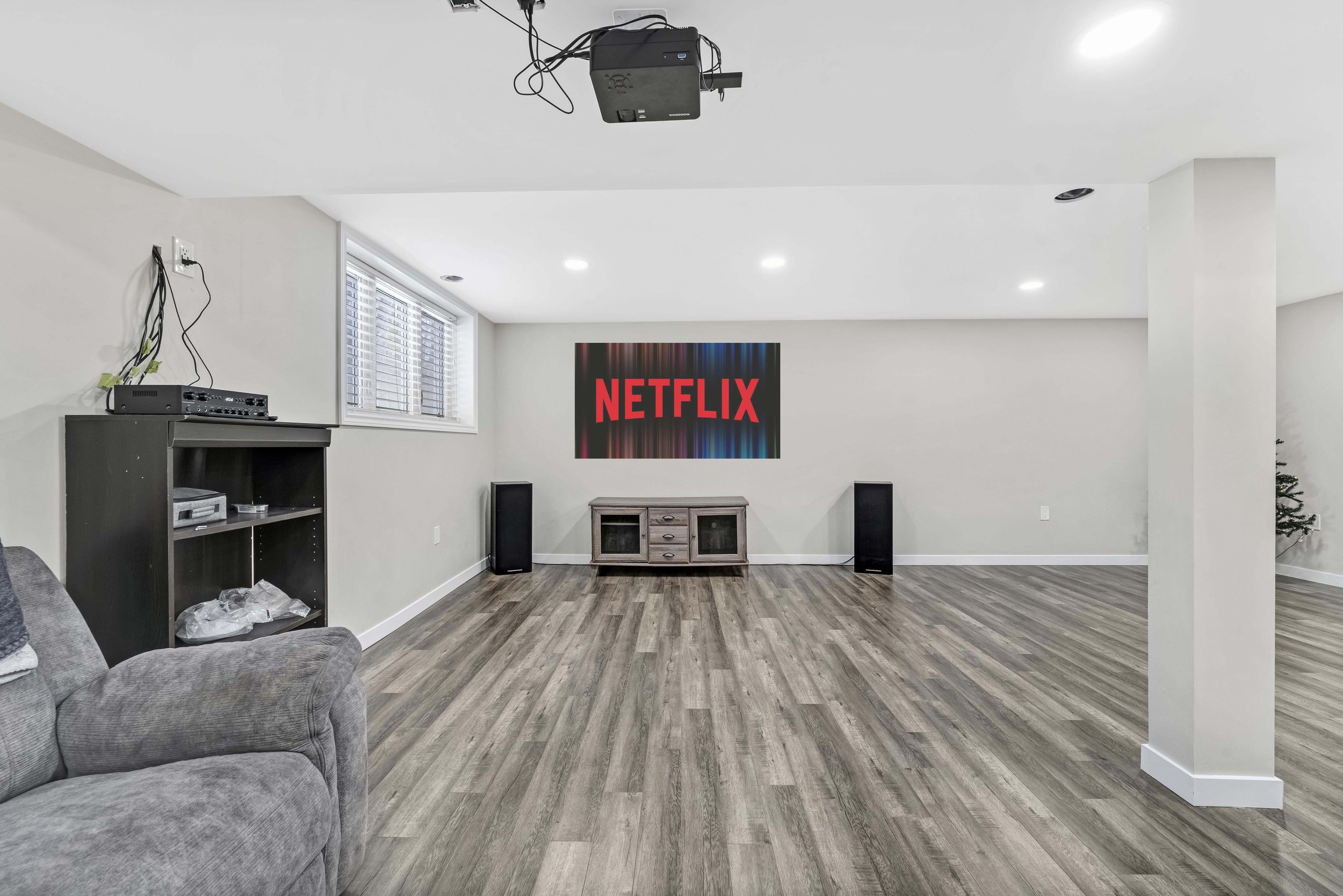 Ottawa Real Estate Photography  Living room with a large flat screen TV displaying the Netflix logo, speakers on either side, a small wooden TV stand, a gray couch, a window with blinds, and a corner Christmas tree.