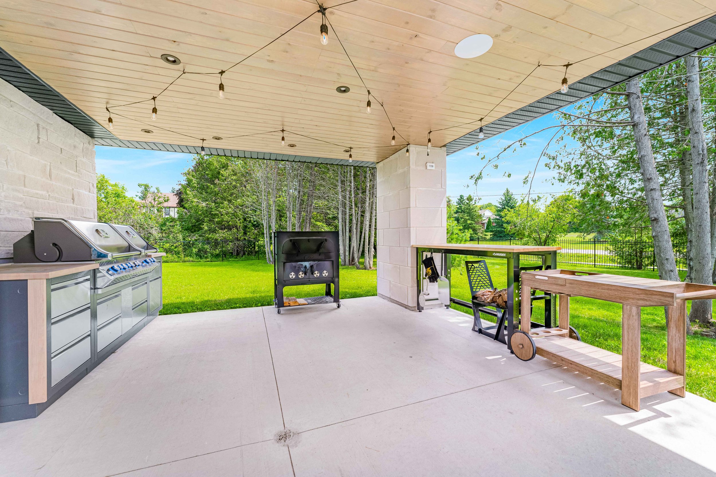 Outdoor covered patio with BBQ grill, string lights, wooden table, and lush green lawn.