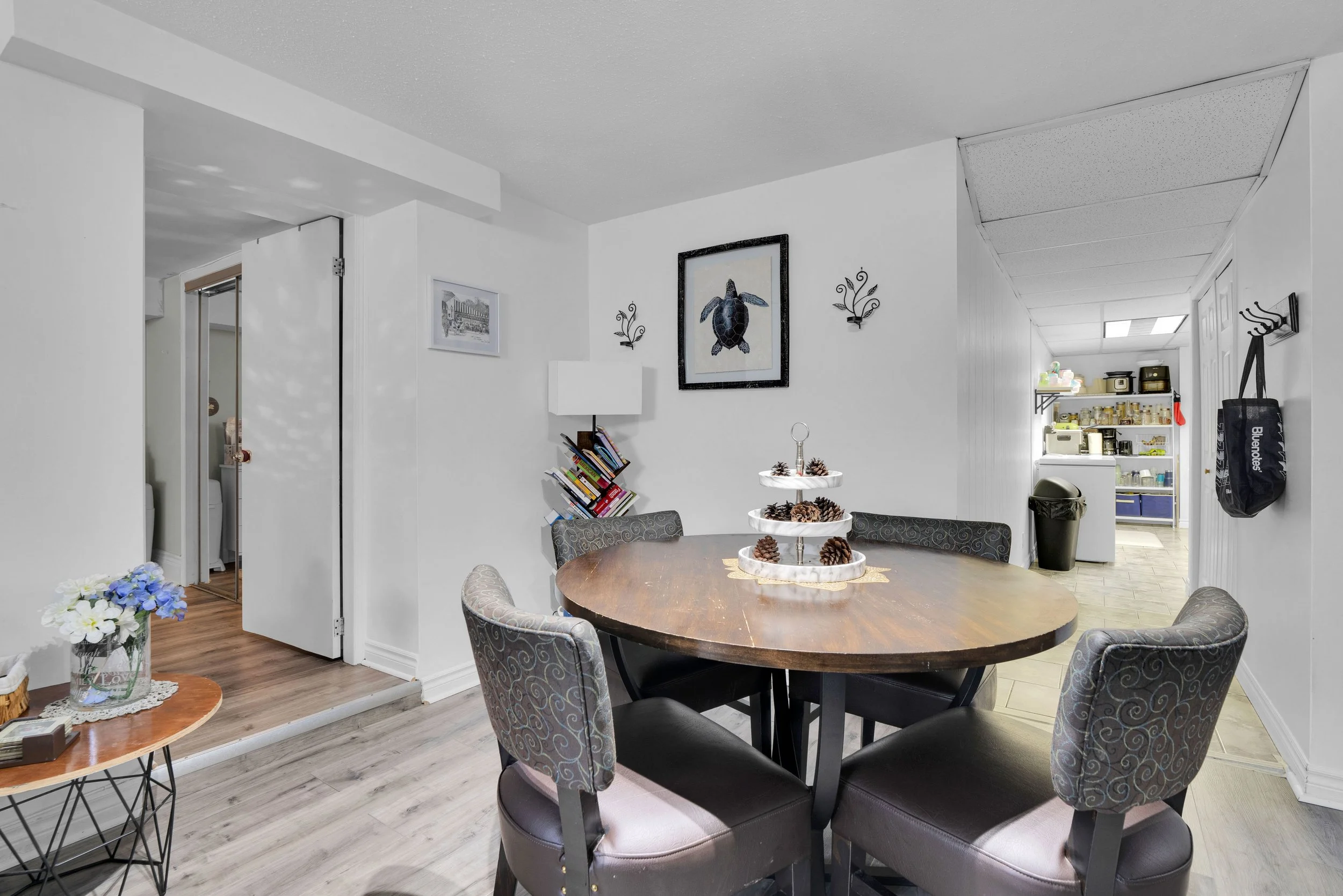 A dining room with a round wooden table surrounded by four upholstered chairs with patterned fabric. A multi-tiered tray with pinecones sits on the table. The walls are painted white and adorned with framed artwork and decorative wall sconces. A smal