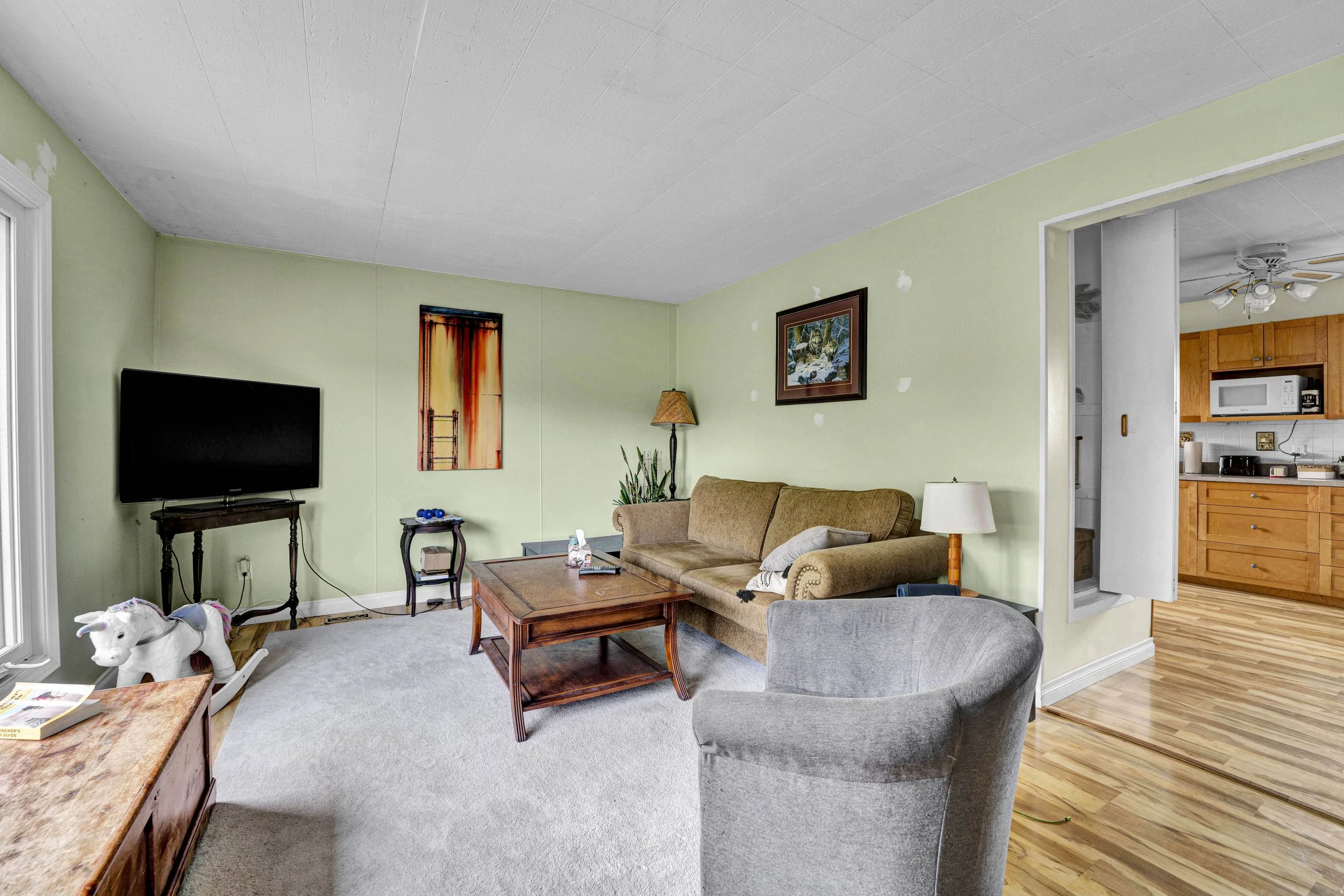 Living room with light green walls, beige sofa, gray armchair, wooden coffee table, floor lamp, wall art, television, and a child's rocking horse.