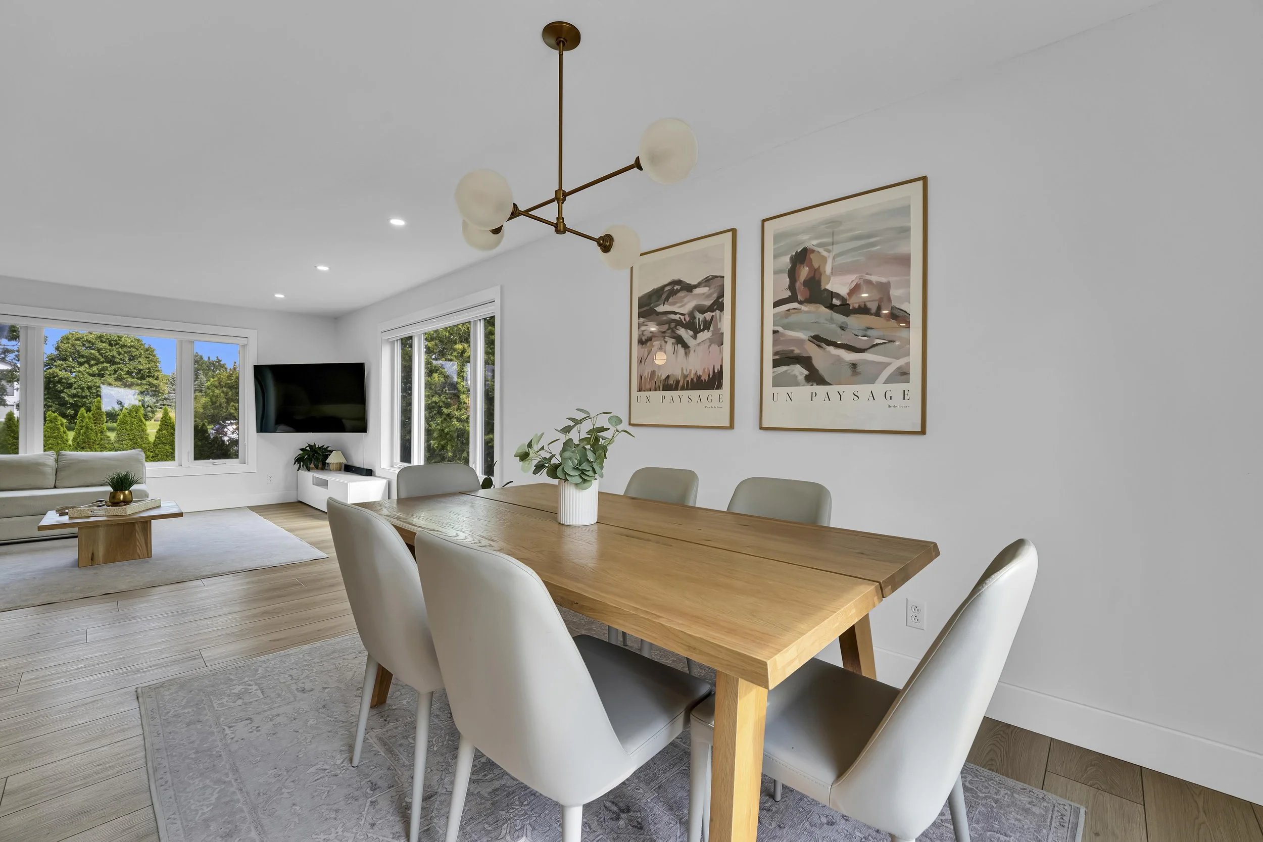 A modern dining room with a wooden dining table, six white chairs, a large window showing a green yard, and a hanging ceiling light fixture with five frosted glass globes. There are two framed landscape art prints on the wall, a potted plant on the t