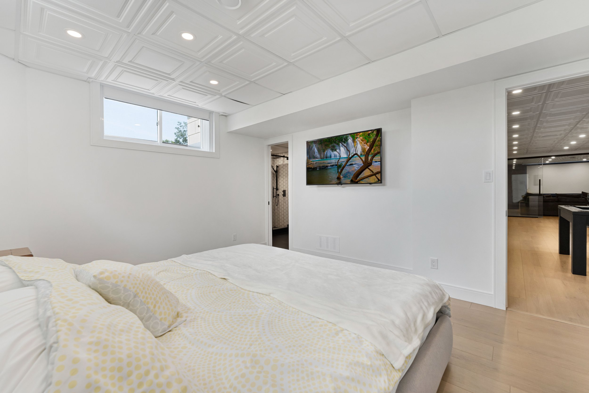 Bedroom with white walls, large bed, patterned bedding, wall-mounted TV, doorway leading to bathroom and another room, recessed lighting, and window with outdoor view.