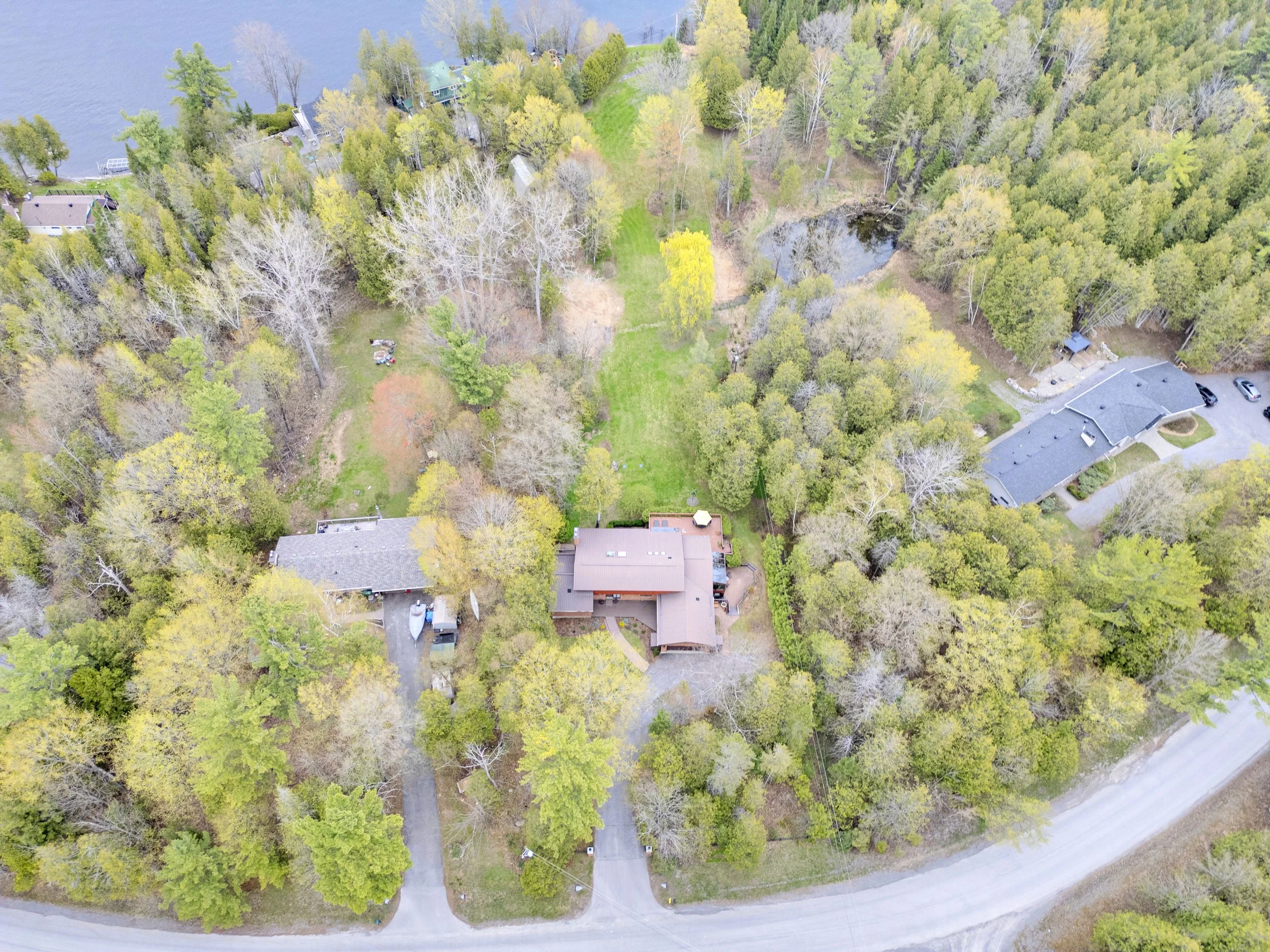 Ottawa Real Estate Media Services  An aerial view of a residential area surrounded by trees and a body of water. Houses are nestled among dense foliage with a small pond visible near the center.