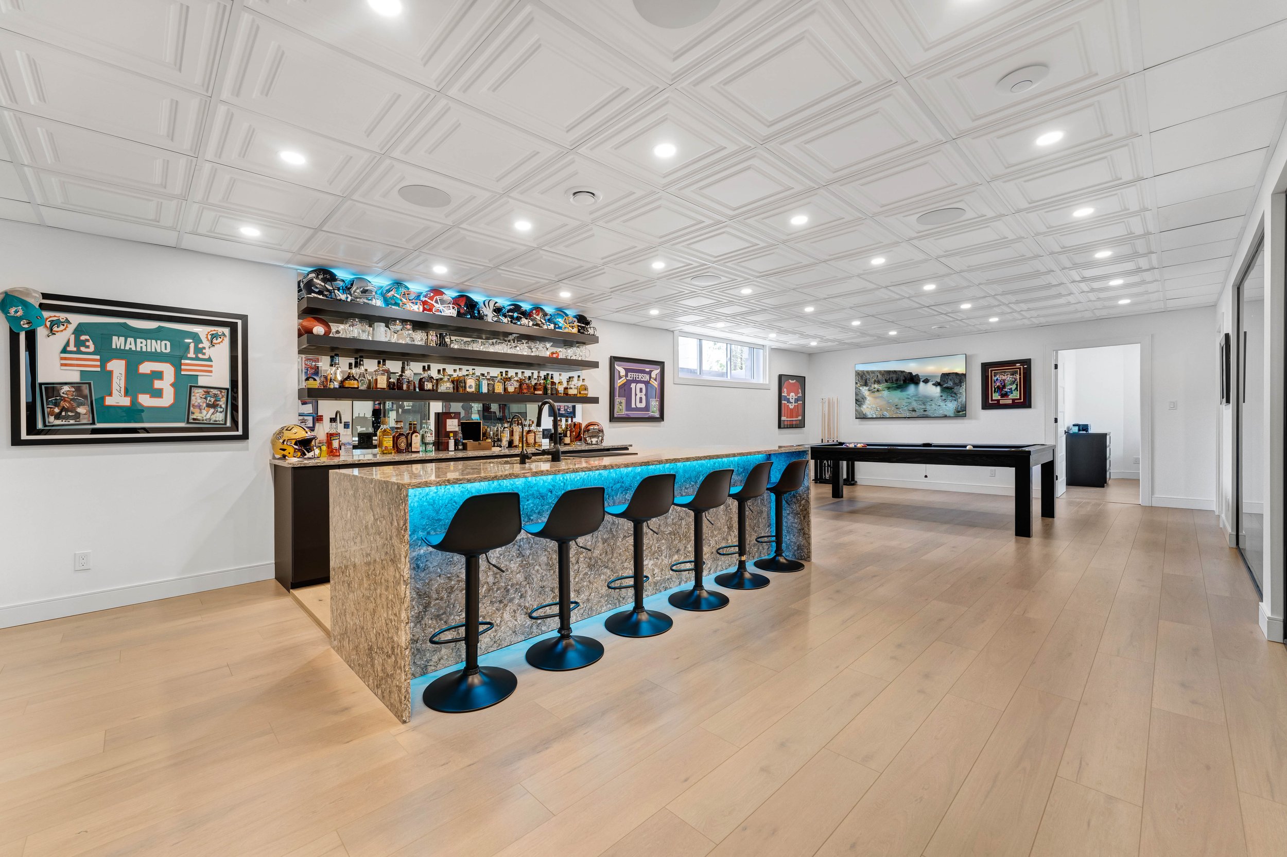 A modern home bar and game room with a bar counter, high chairs, sports memorabilia, framed sports jerseys, a collection of drinks, and a pool table.