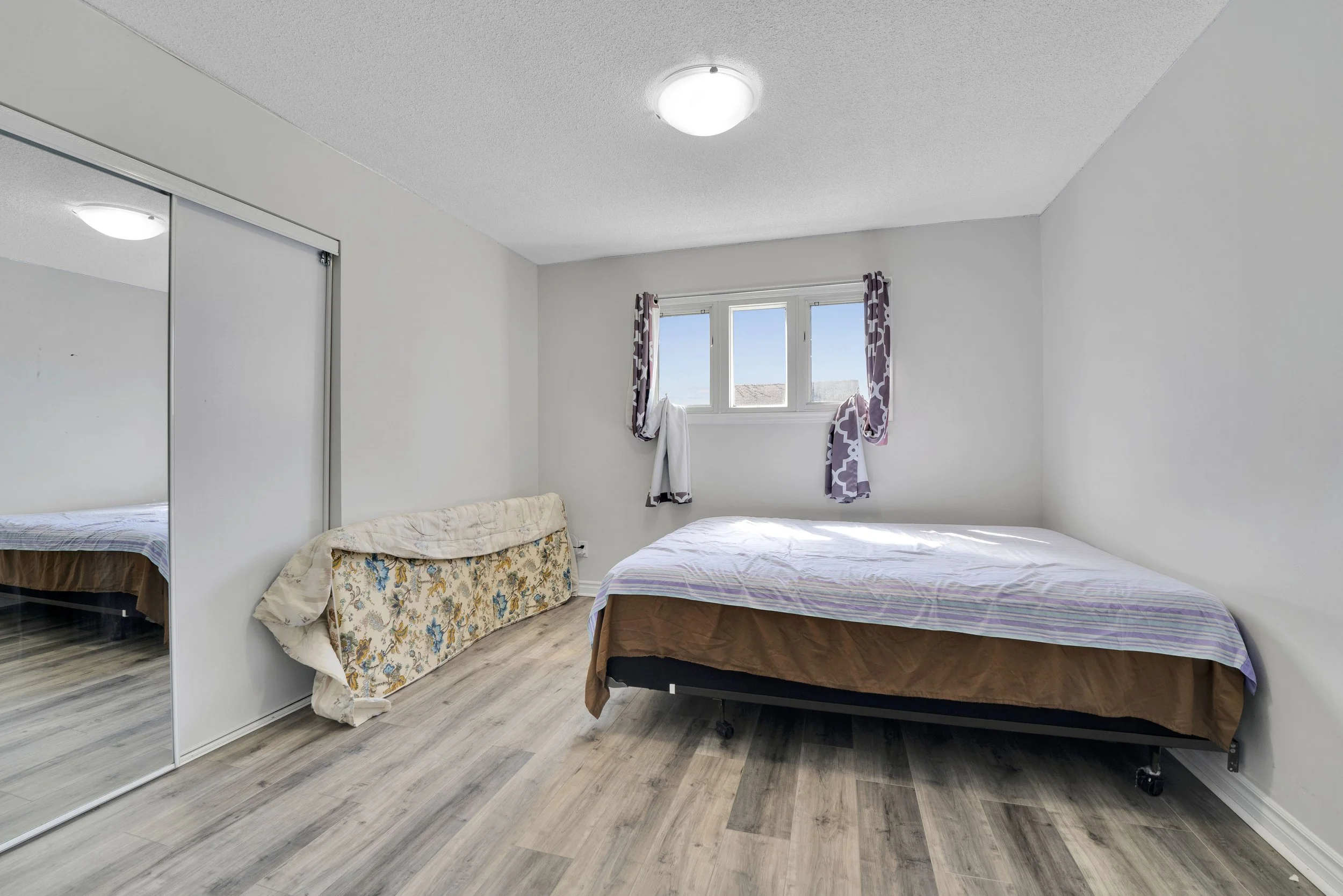 Ottawa Real Estate Media Services  A bedroom with white walls, a window with purple and white patterned curtains, a bed with a brown and purple bedspread, a floral-patterned couch, and a closet with sliding mirror doors.