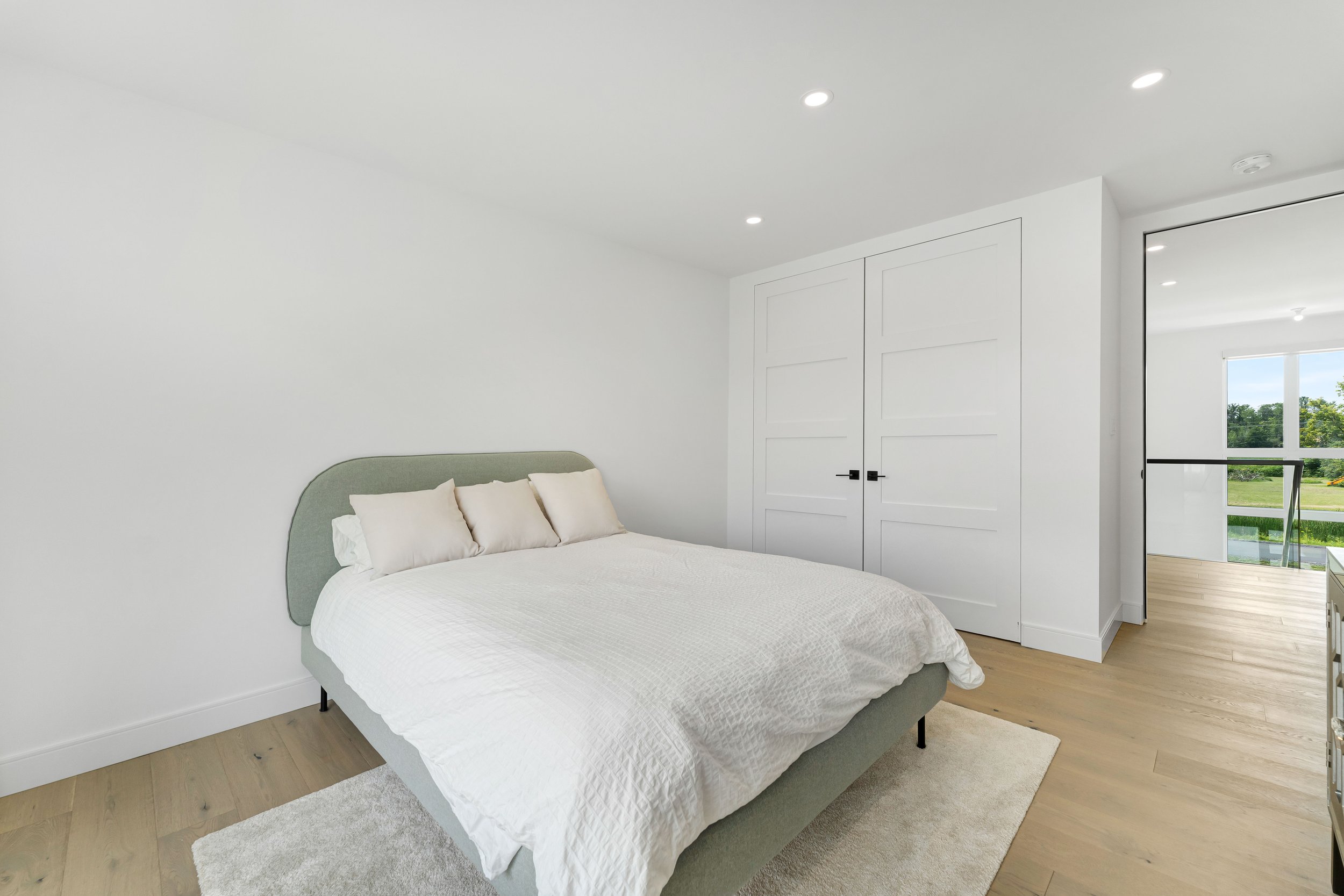 Minimalist bedroom with a gray bed, white bedding, and matching pillows. Light wood flooring, white walls, and a built-in closet with double doors. Large window with views outside.