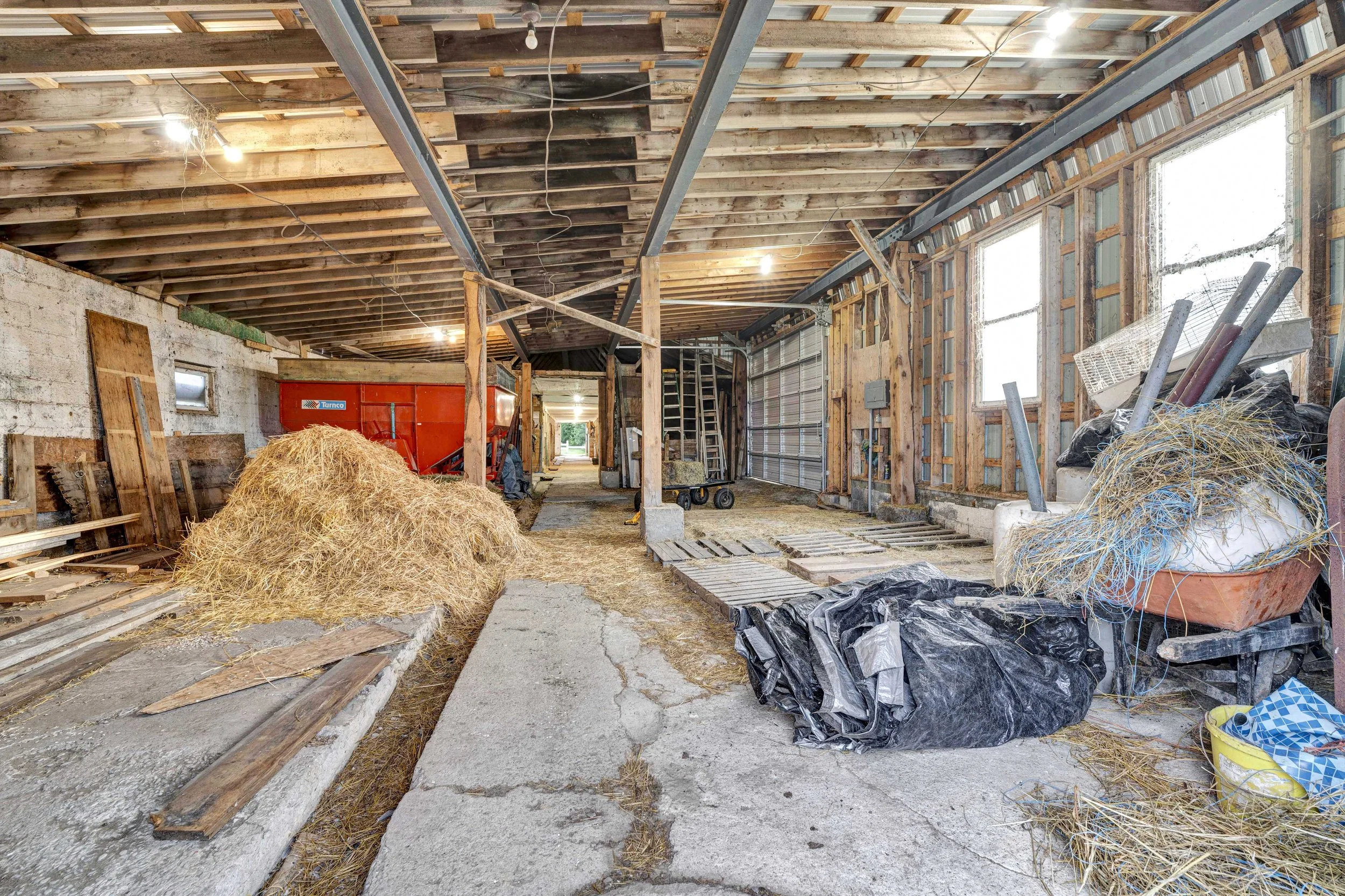 This is an unfinished barn or shed with exposed wooden beams, construction materials, and farm equipment. Hay bales, a wheelbarrow, and pipes are on the floor, with windows on the right, and a garage door at the back. Temporary lighting and scaffoldi