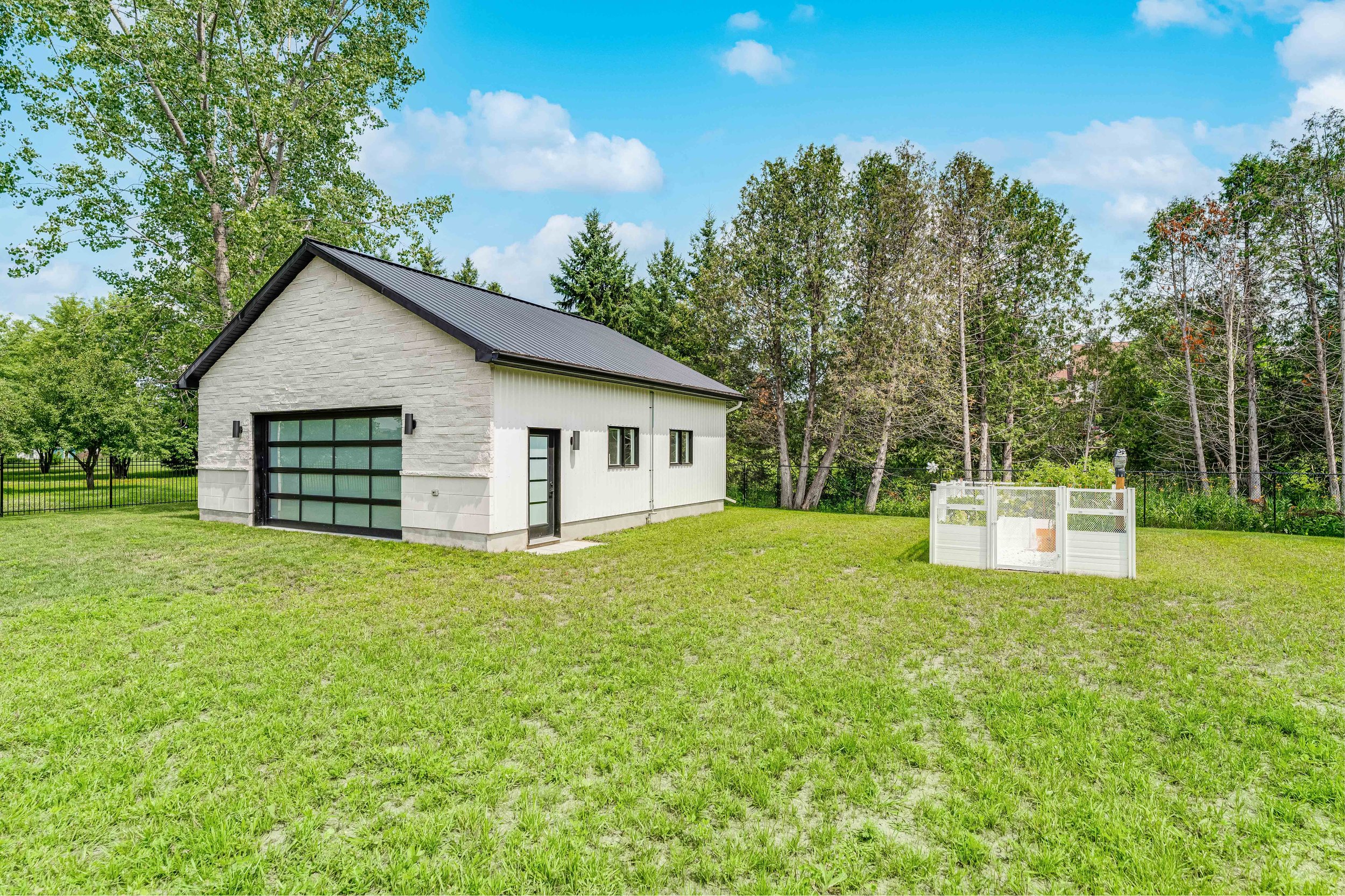 A modern white garage with a black metal roof situated on a grassy lawn, surrounded by trees. The garage has a glass panel door and side entrance, with a small fenced area nearby under a clear blue sky.