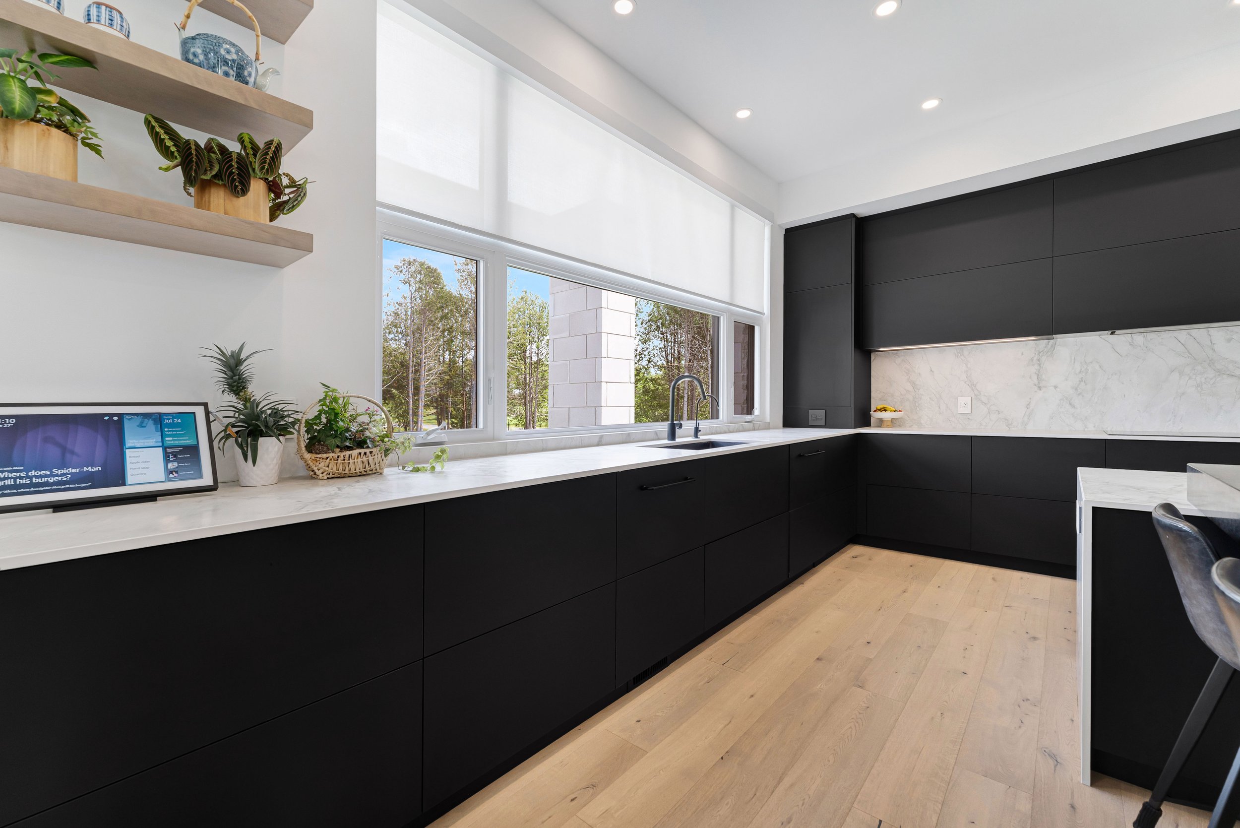 Modern kitchen with black cabinets, white marble countertops, light wood flooring, and a large window. Features open shelves with plants and decorative items, and a digital device on the counter.