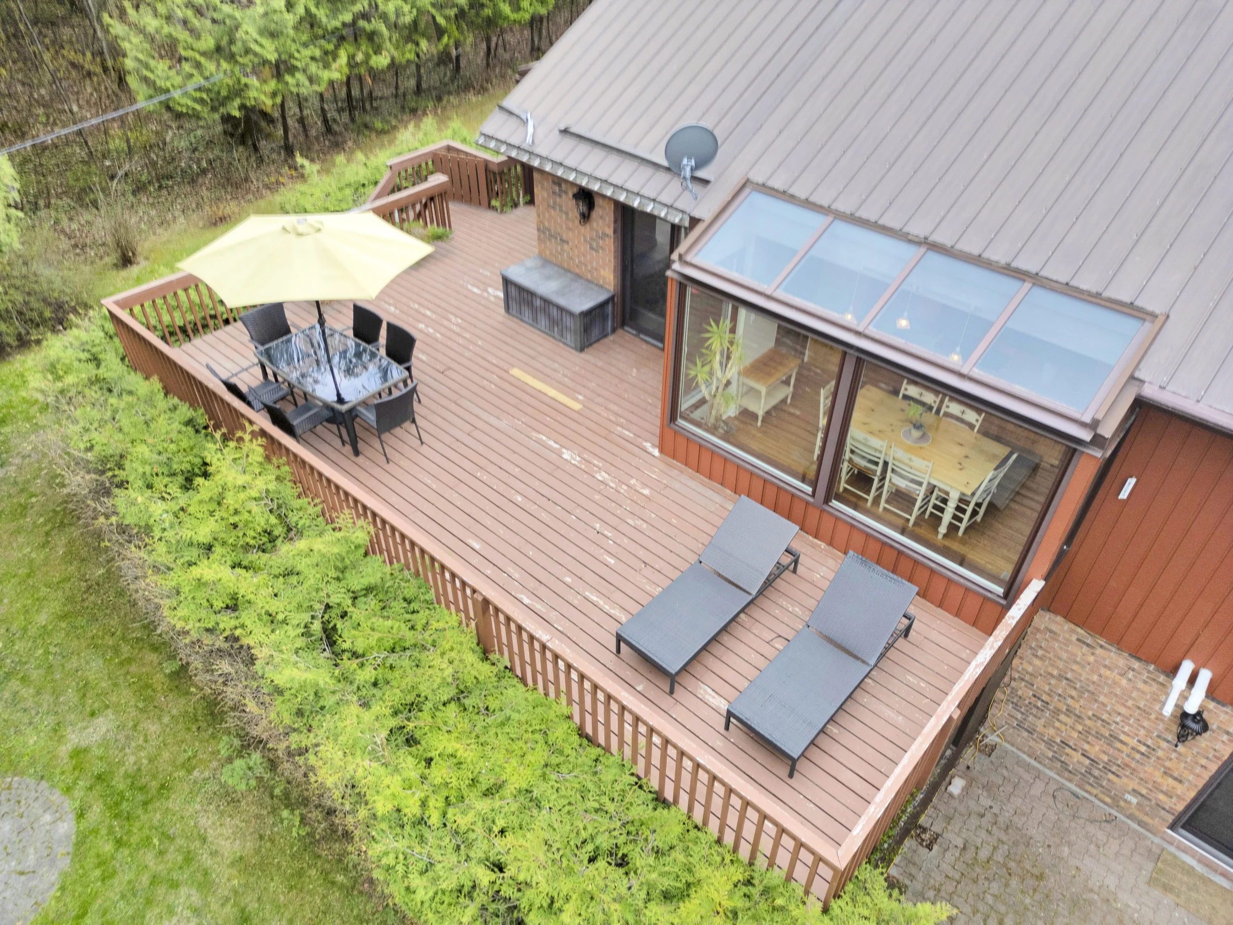 Ottawa Real Estate Media Services  An aerial view of a wooden deck attached to a house with a large window and outdoor dining area, surrounded by trees and greenery.