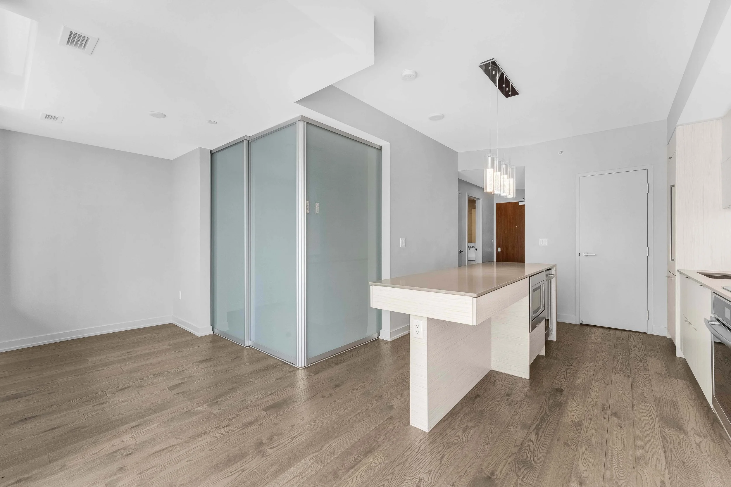 NV3D Ottawa Real Estate Photography Empty kitchen and living area with wooden flooring, modern lighting fixtures, a glass shower enclosure, and minimal white and light beige walls.