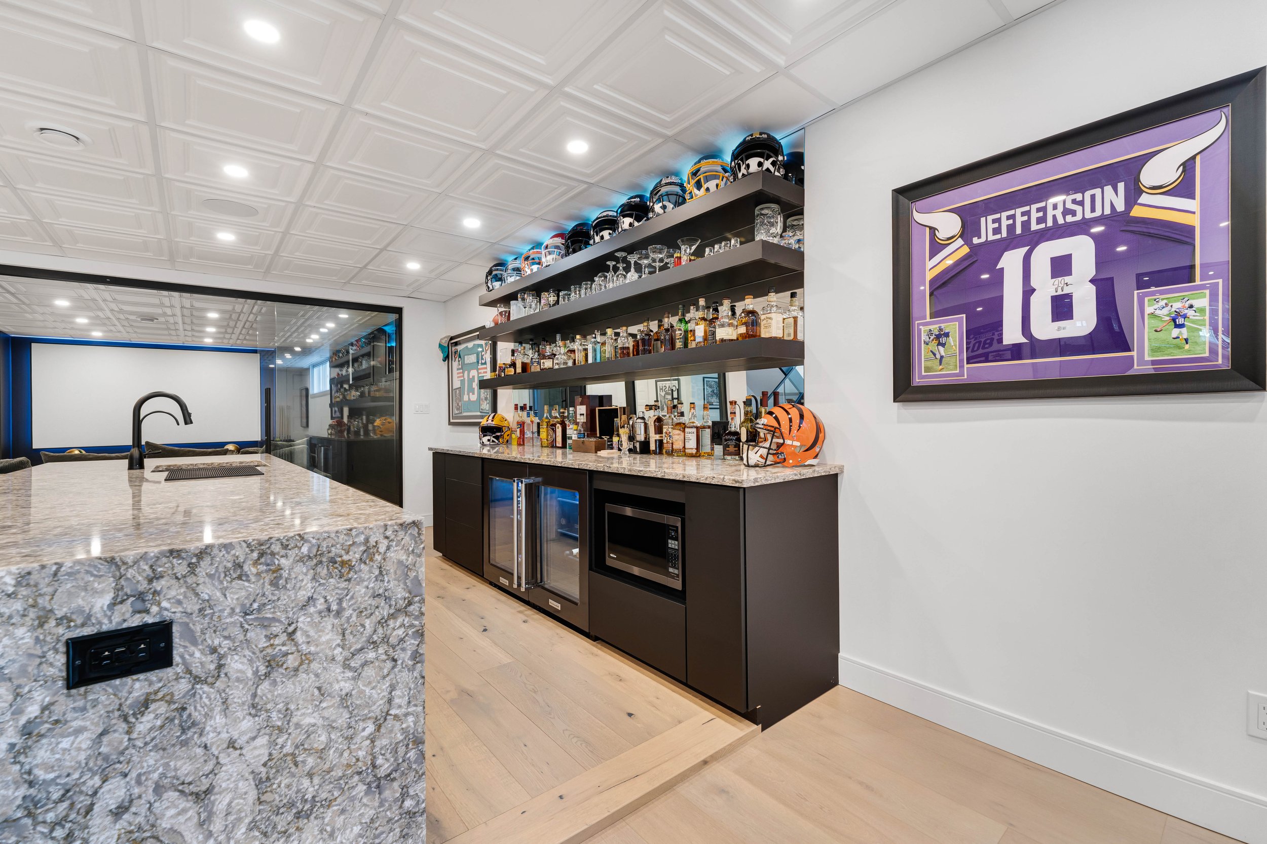 Modern home bar with sports memorabilia, display shelves with bottles and glasses, framed jersey, and football helmets. Granite countertop with sink and appliances.