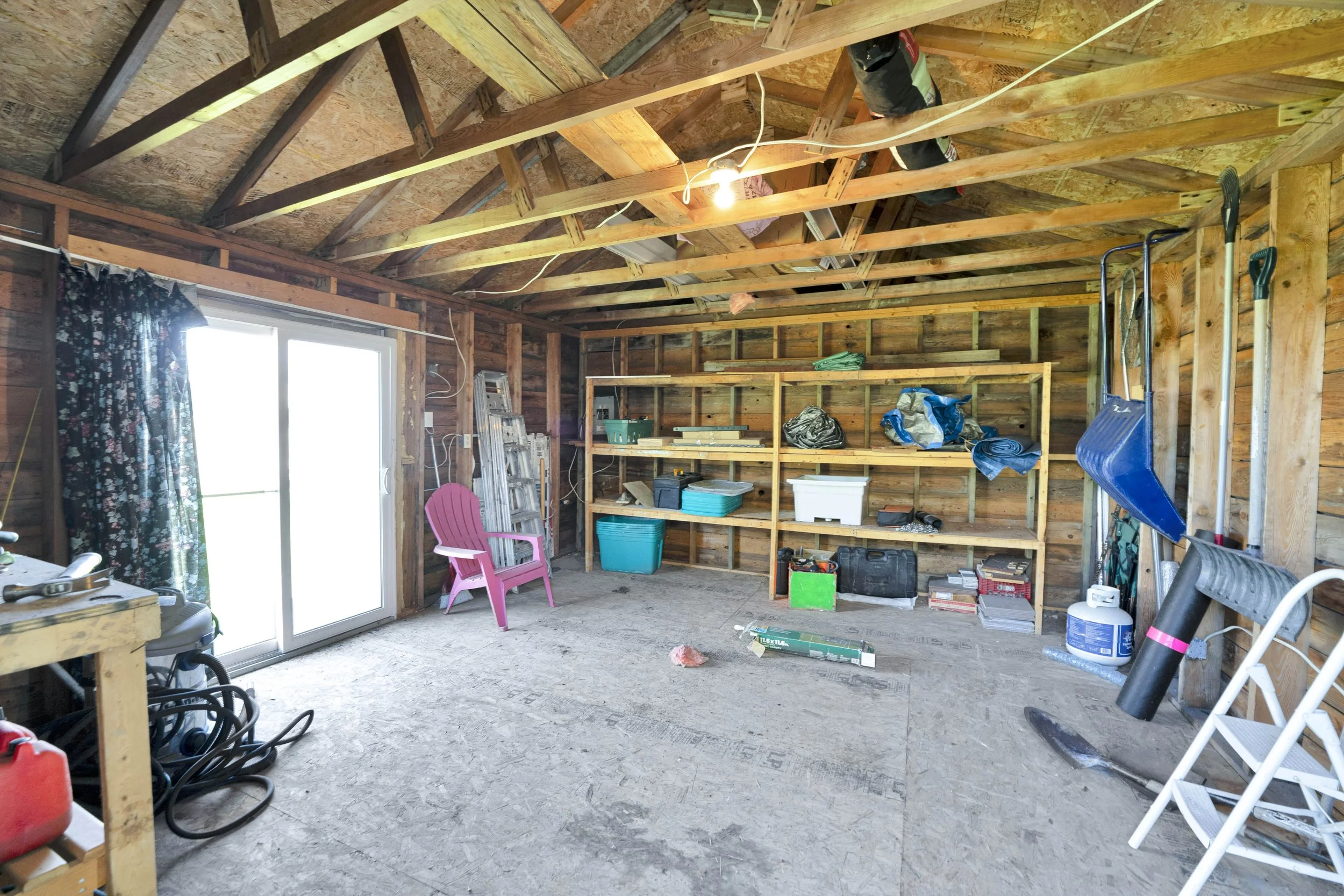 Ottawa Real Estate Media Services  A room under construction or renovation with exposed wooden framing and insulation, a sliding glass door with a floral curtain, various tools, storage bins, and a pink plastic chair.