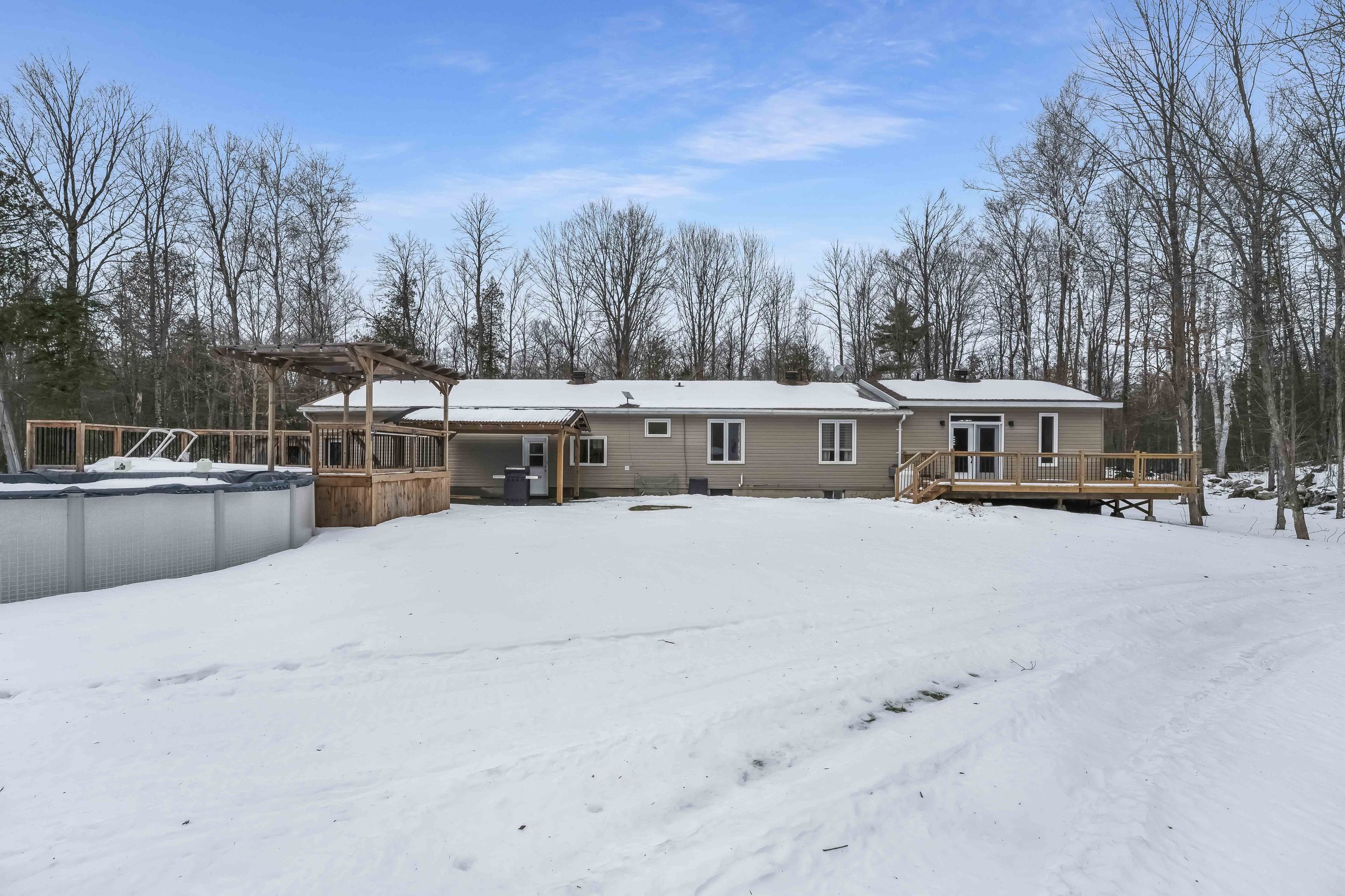 Backyard with snow-covered ground, wooden deck, above-ground pool, house with multiple windows and doors, fenced area, leafless trees, and a clear blue sky.