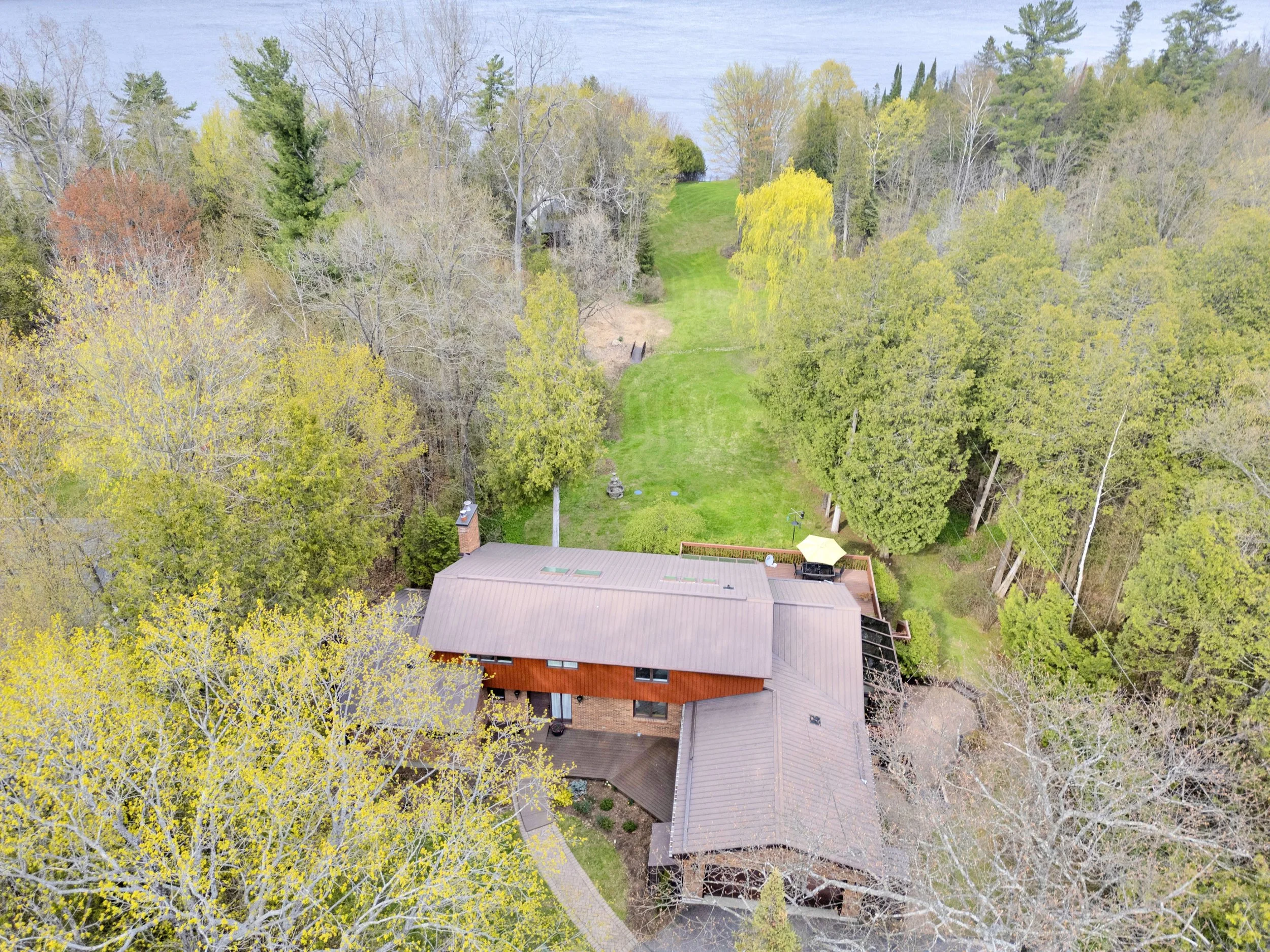 Ottawa Real Estate Media Services  An aerial view of a house surrounded by trees, with a grassy yard, near a lake.