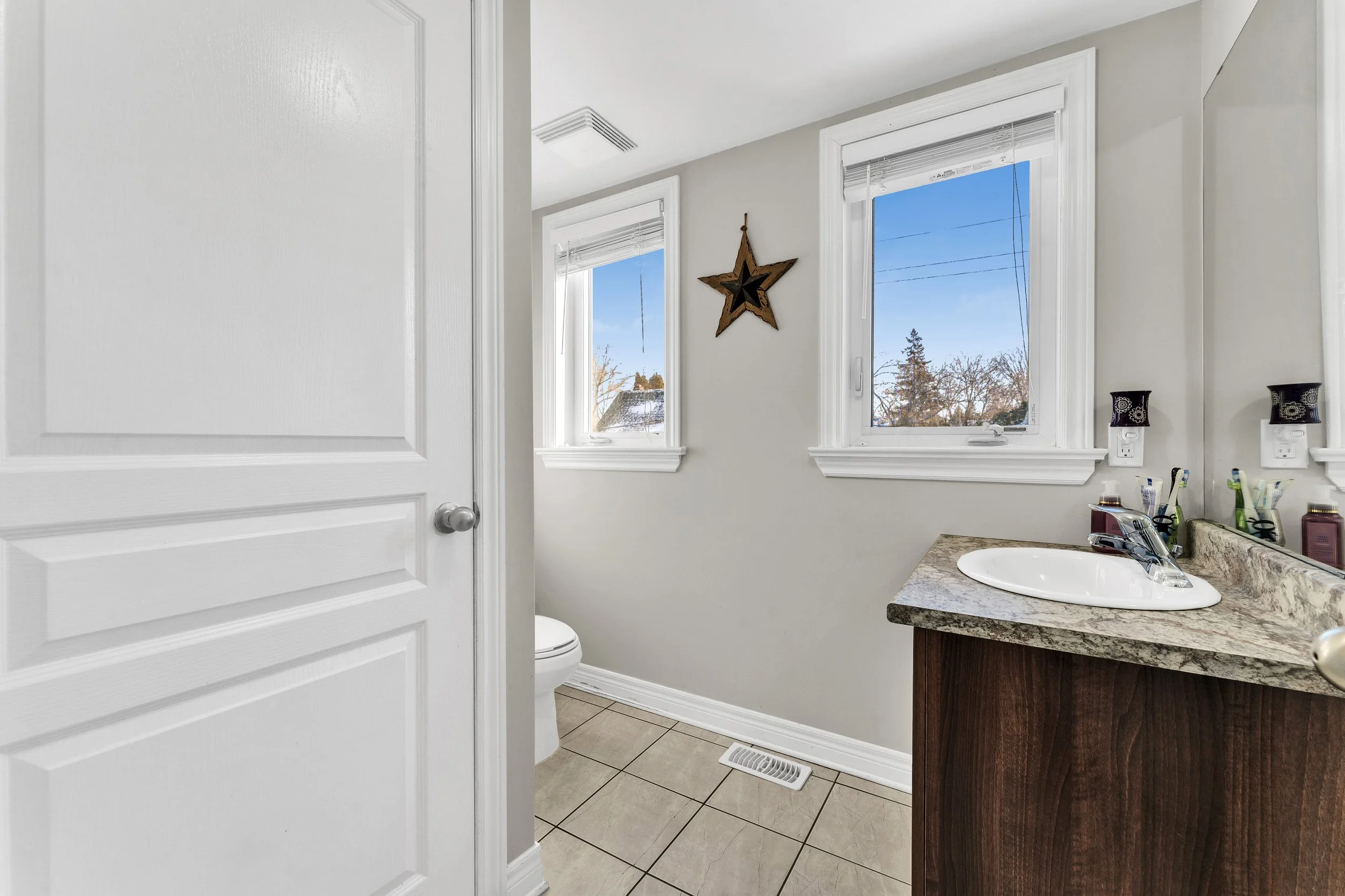 Ottawa Real Estate Photography  Bathroom with two windows showing a blue sky and trees, a star wall decoration, a toilet, and a dark wood vanity with a granite countertop and a sink.