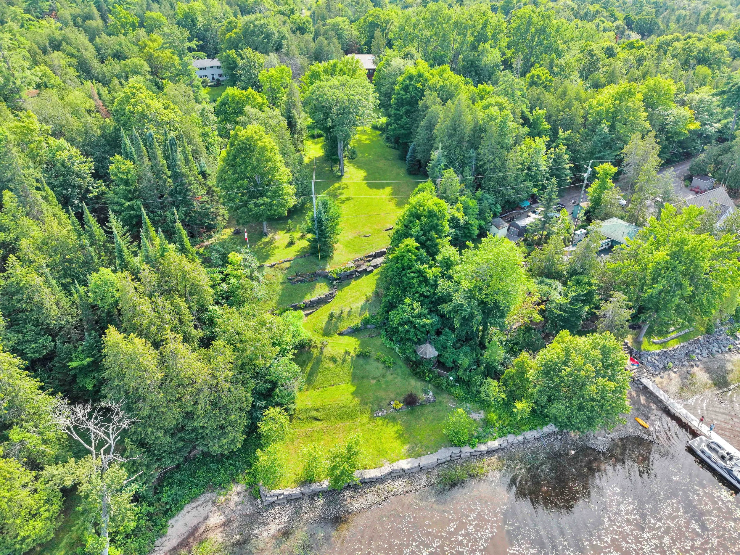 Ottawa Real Estate Photographer Aerial view of a lush, green forest with a small clearing, a river at the bottom, and a few houses and structures among the trees.