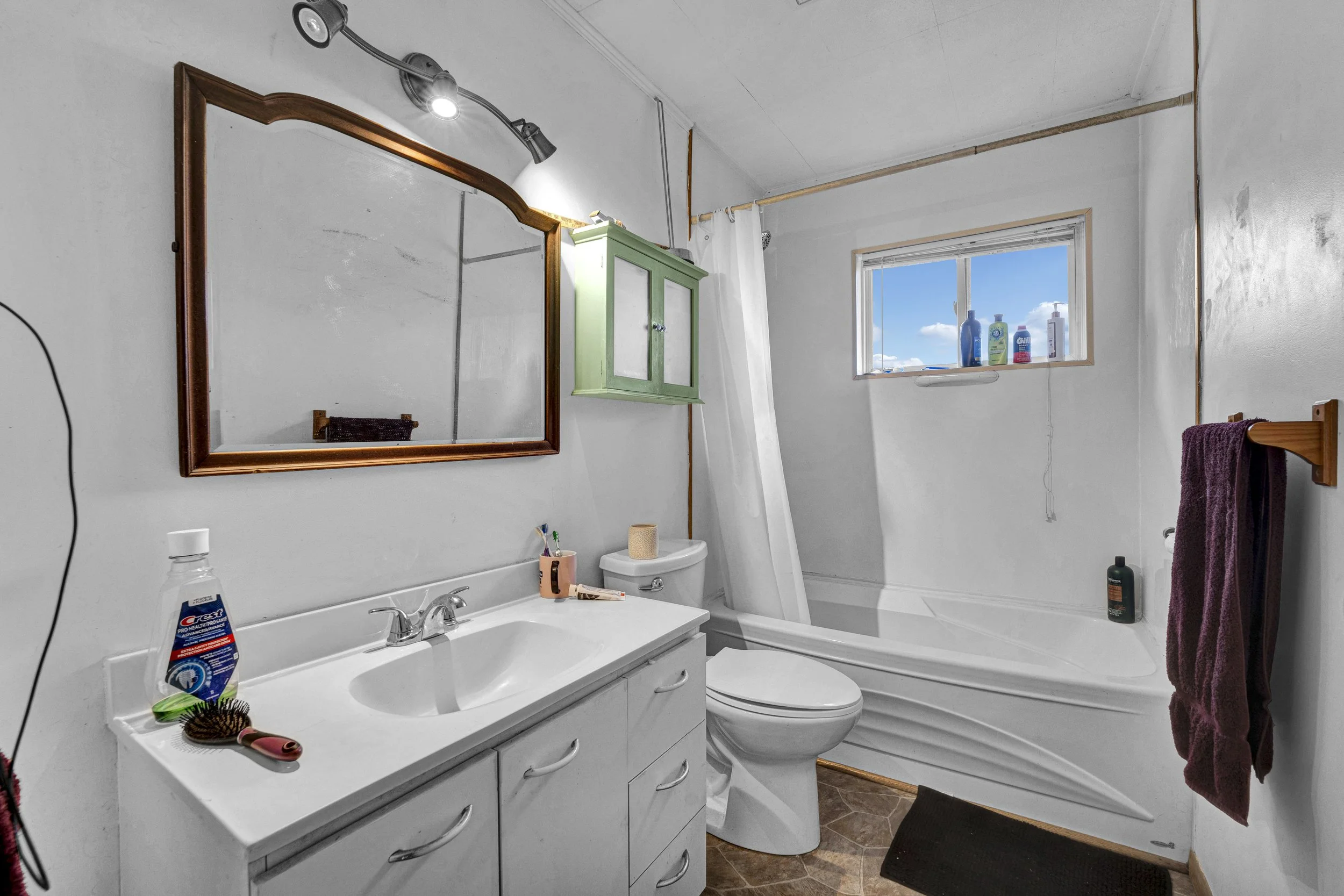A small bathroom with a white vanity, mirror, toilet, and bathtub. The bathroom has a window with a view of blue sky and clouds. Items like shampoo, soap, and cleaning supplies are on the windowsill and around the room. Purple towel hanging on the wa
