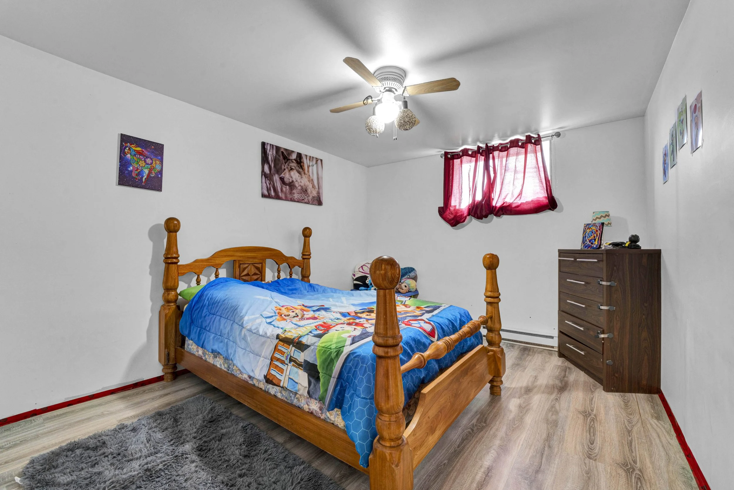 A wooden bed with a colorful cartoon-themed bedspread in a bedroom with white walls, red curtains, artwork of a wolf and a unicorn on the walls, and a wooden dresser, with a dark gray rug on light-colored flooring.