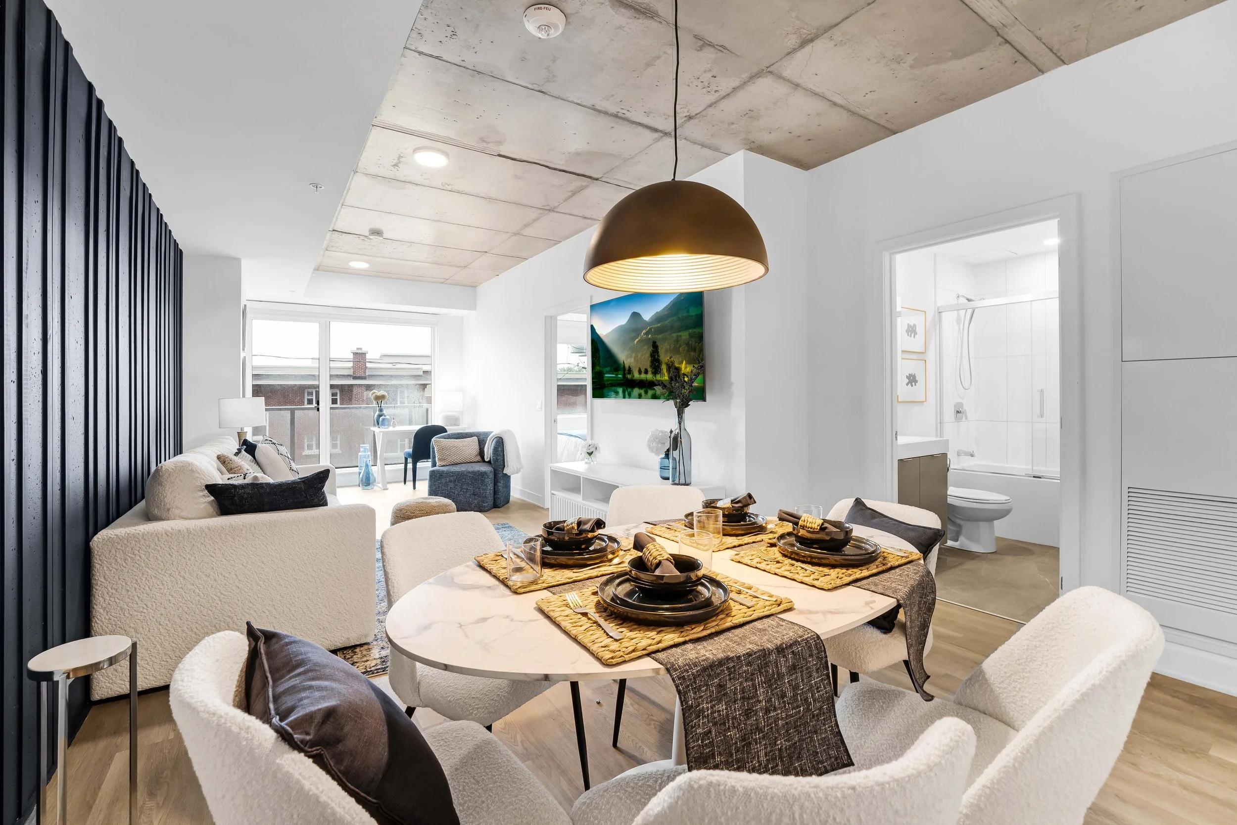 Modern apartment living room and dining area with white walls, wooden floors, and a concrete ceiling. The dining table is set with black plates, gold cutlery, and woven placemats. The living room has a cream-colored sofa with decorative pillows, a bl