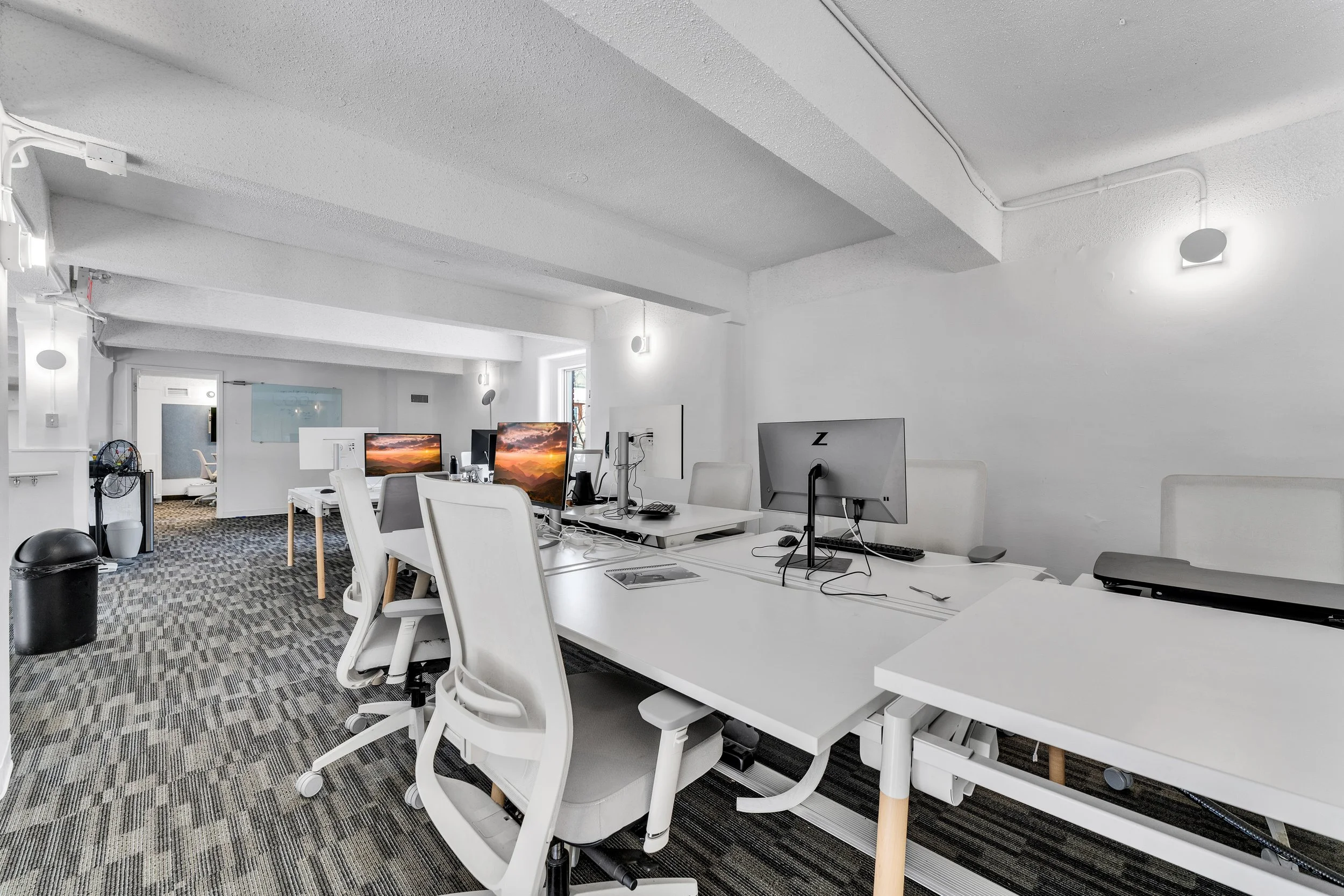 A modern, minimalist office space with white desks, ergonomic office chairs, multiple computer monitors, and a neutral-colored carpet. The room is well-lit with natural and artificial lighting, and there is a door and some office equipment in the bac