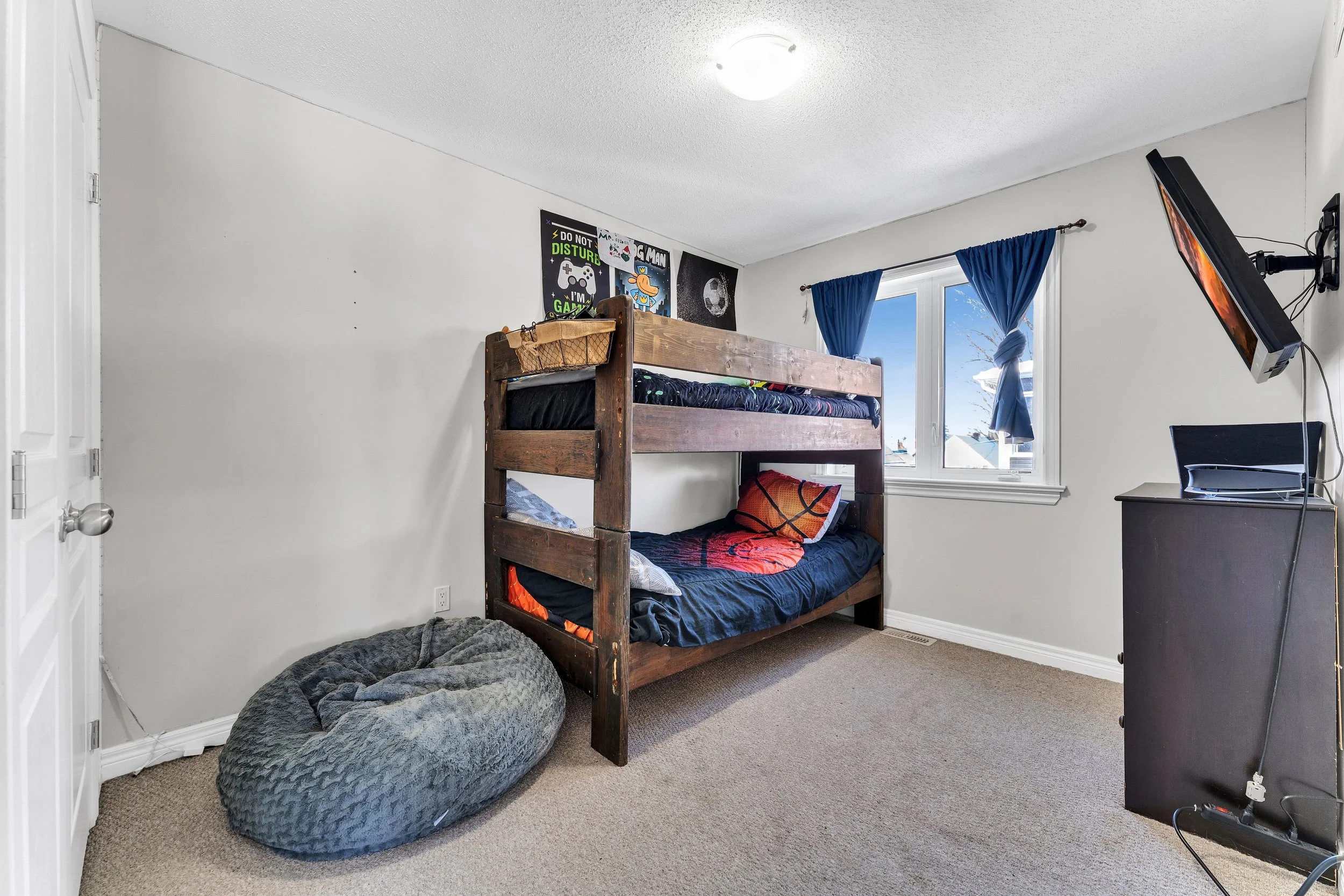 A bedroom with a bunk bed, a bean bag chair, a window with blue curtains, and a television mounted on the wall.