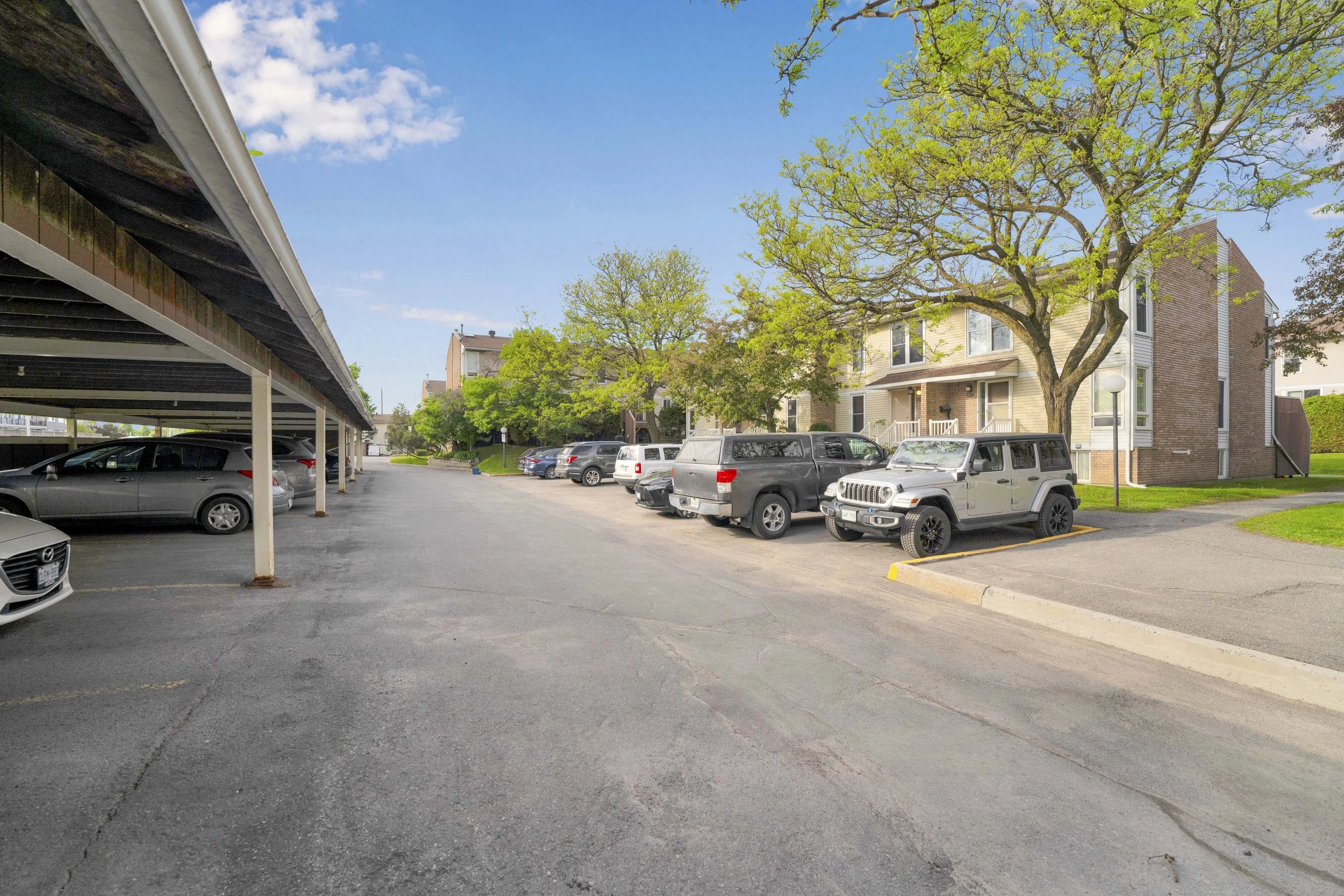 A parking lot with cars parked under a carport on the left. Residential buildings with trees and a partly cloudy sky in the background.
