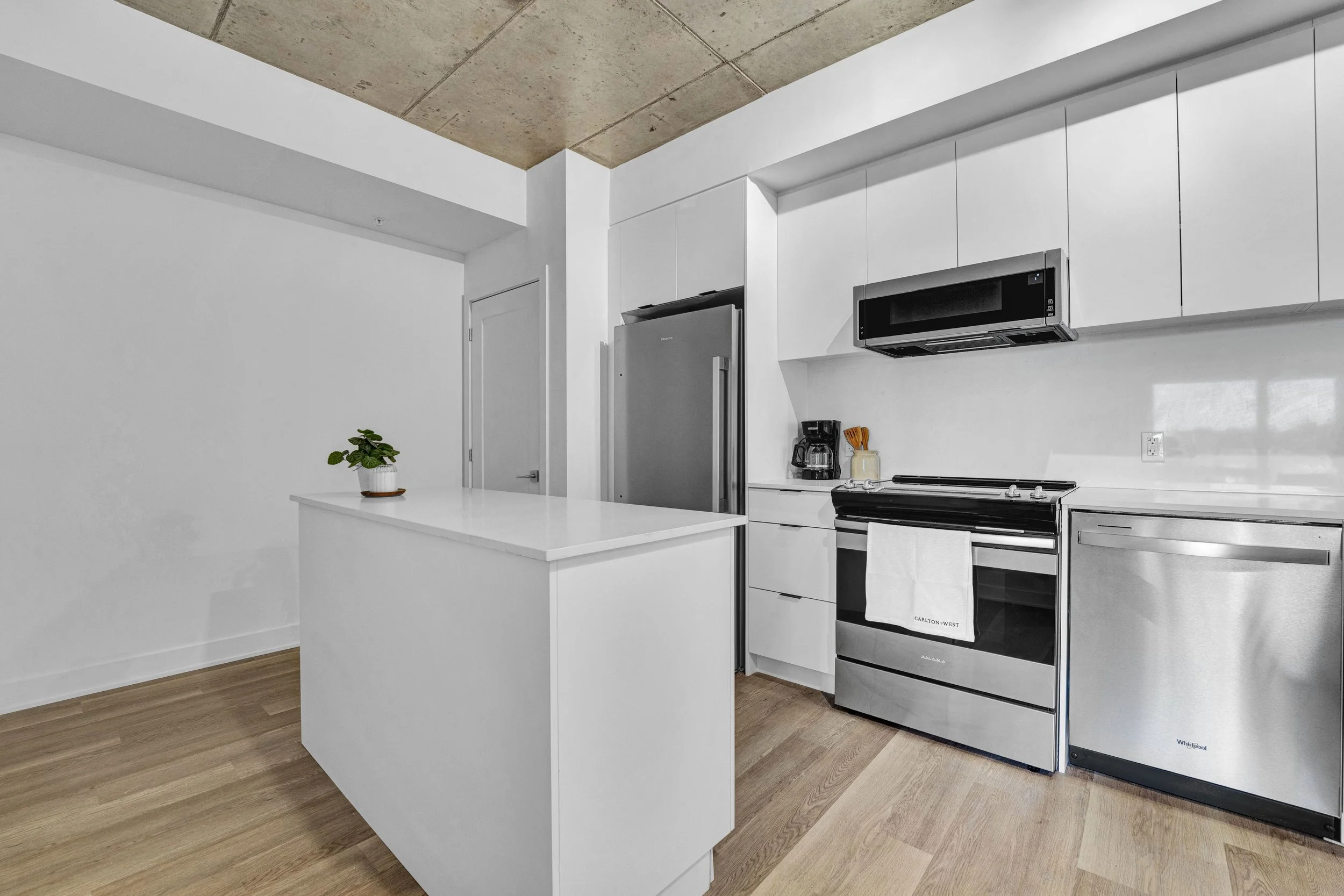 Ottawa Real Estate Media Services  Modern kitchen with white cabinets, stainless steel refrigerator, black microwave, electric stove, coffee maker, and a small potted plant on the white island, with wooden flooring and a concrete ceiling.