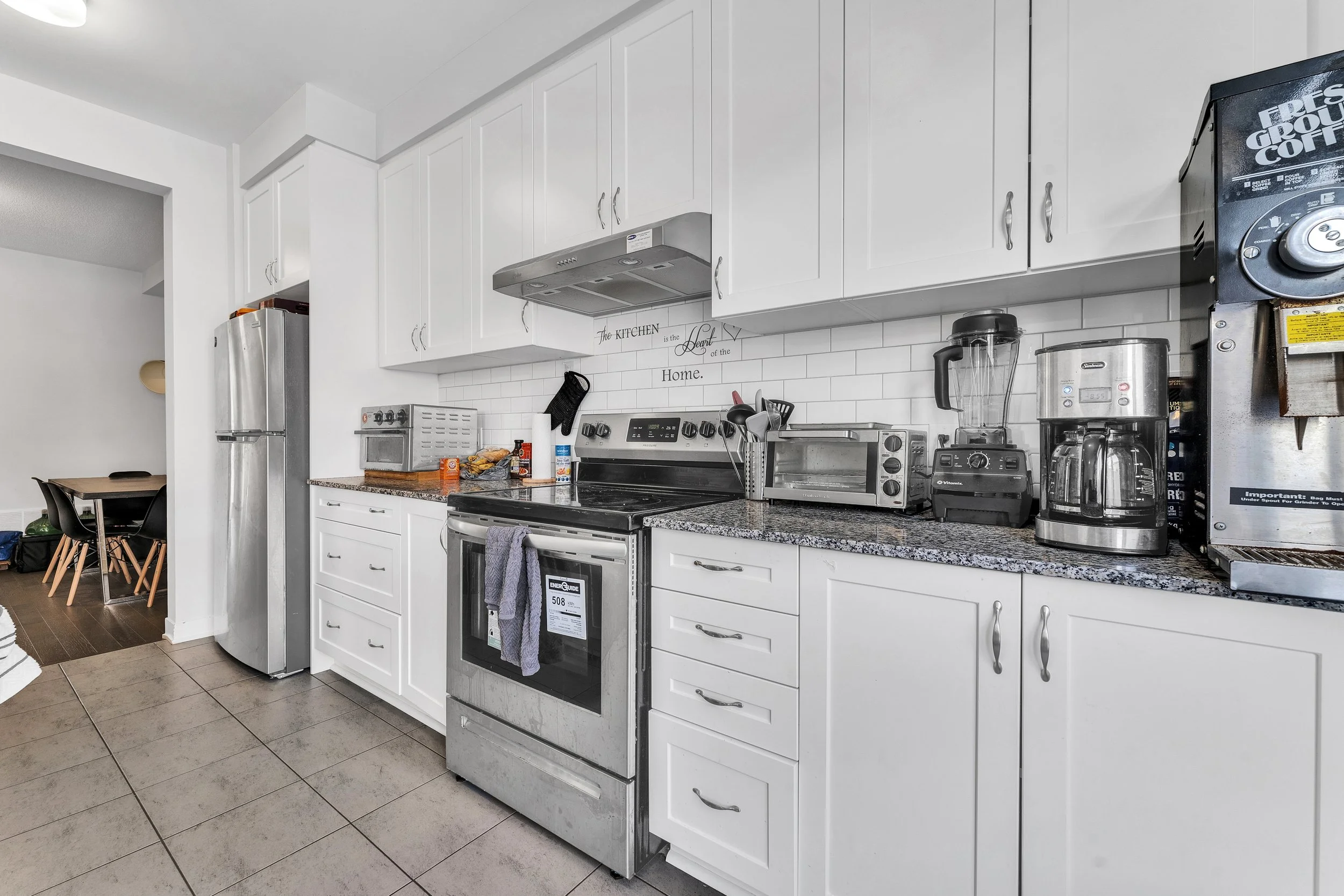 Kitchen with white cabinets, granite countertop, stainless steel appliances including a refrigerator and oven, and various small appliances like a toaster oven, microwave, blender, coffee maker, and a commercial coffee machine on the counter.