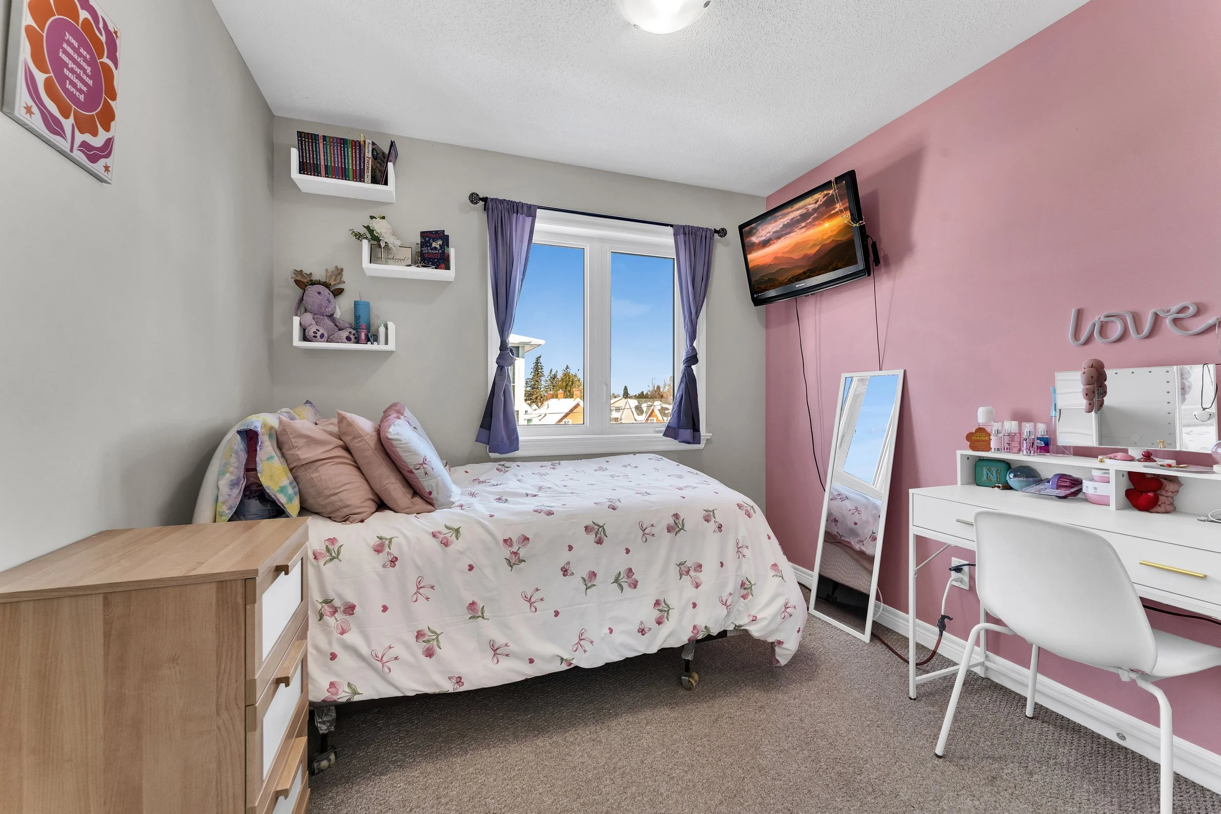 A bedroom with a bed covered in white sheets with pink floral designs and pink pillows, a small wooden dresser on the left, a window with purple curtains, a wall-mounted TV showing a sunset, a mirror on the floor, a white vanity table with a chair, a