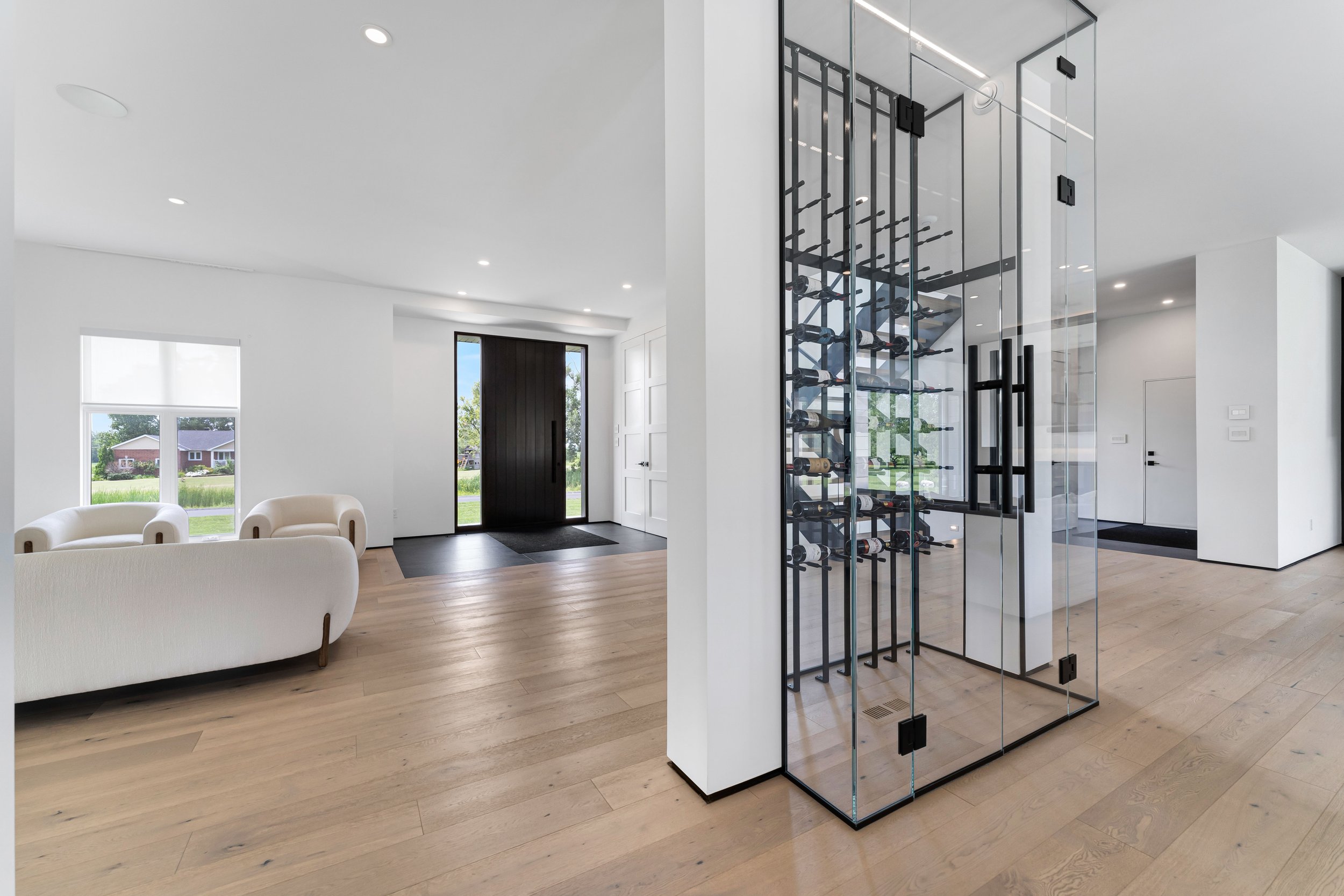 Real estate photography ottawa Modern living space with minimalist design, featuring light wooden flooring, white walls, a seating area with white couches, a glass wine display, and large window with natural light.